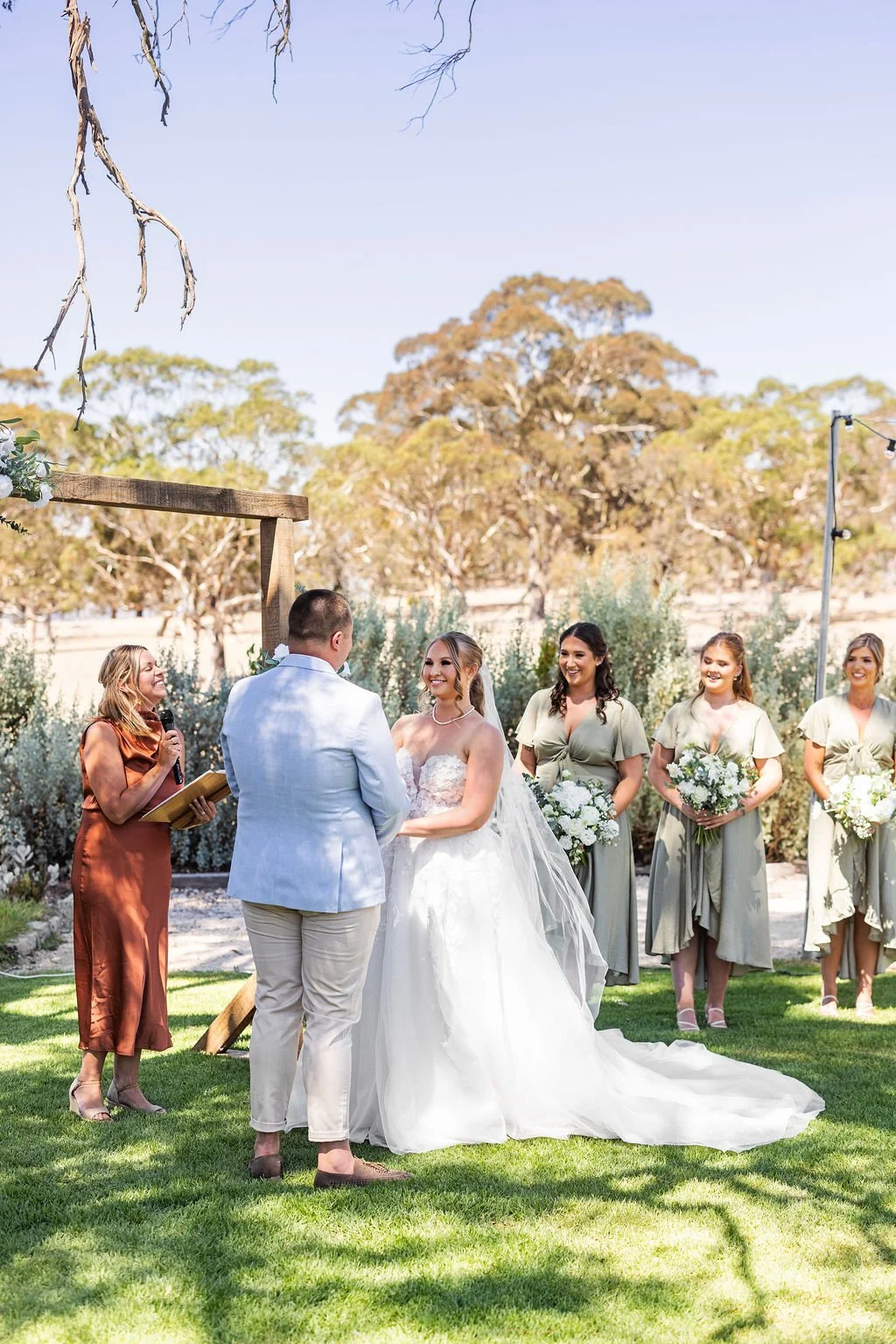 A wedding ceremony taking place outdoors on a sunny day with the bride and groom standing in front of a minister, surrounded by bridesmaids holding bouquets, with trees and clear blue sky in the background.