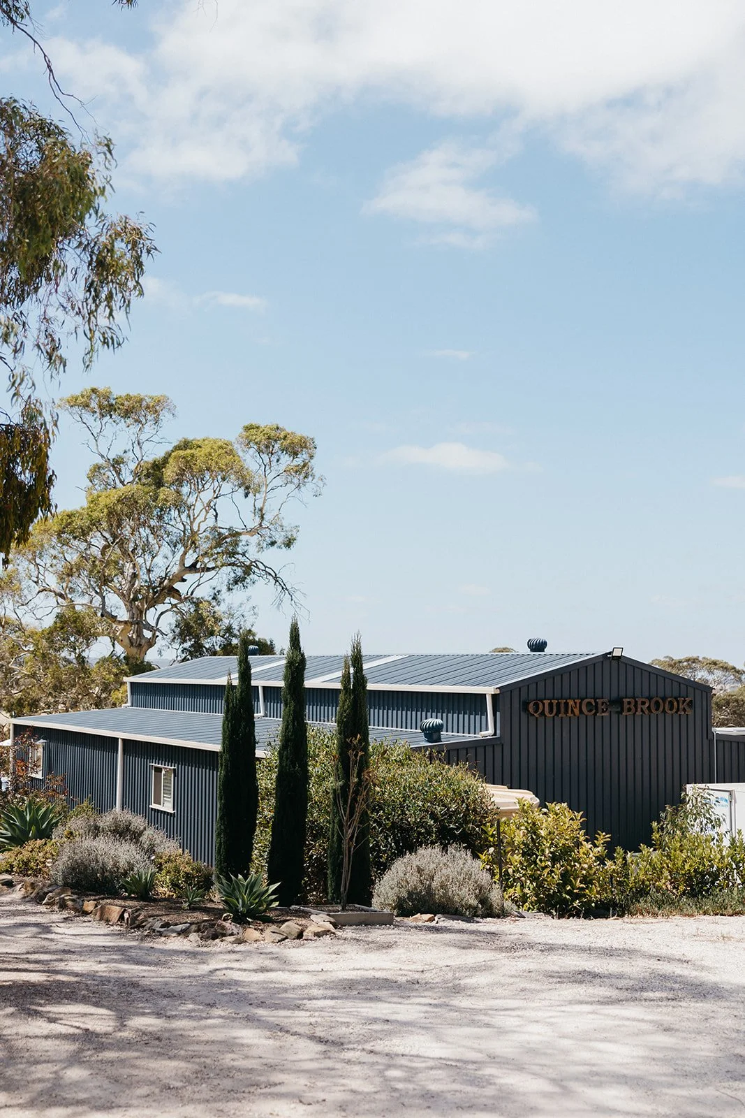 A modern blue building with a metal roof and the sign 'Quince Brook' on it, surrounded by trees and desert vegetation under a partly cloudy sky.