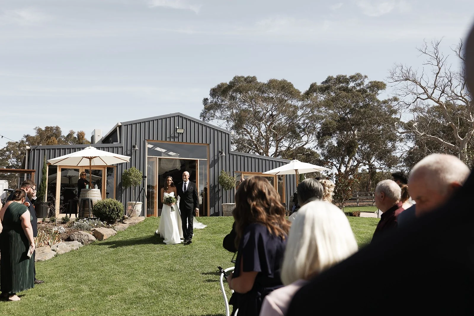 Wedding ceremony outdoors with a bride walking with a man in a suit, guests seated on grass, and a modern building in the background.