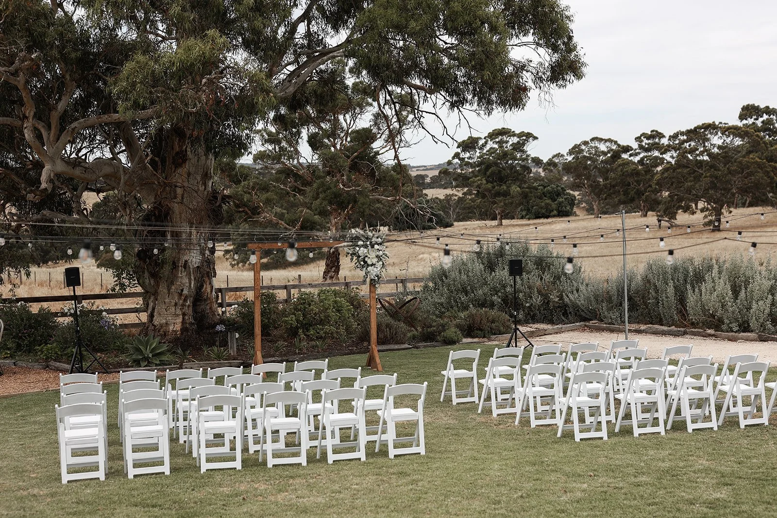Outdoor wedding or event setup with multiple white chairs arranged on a grassy area, large trees and string lights overhead, in a rural landscape with rolling fields in the background.