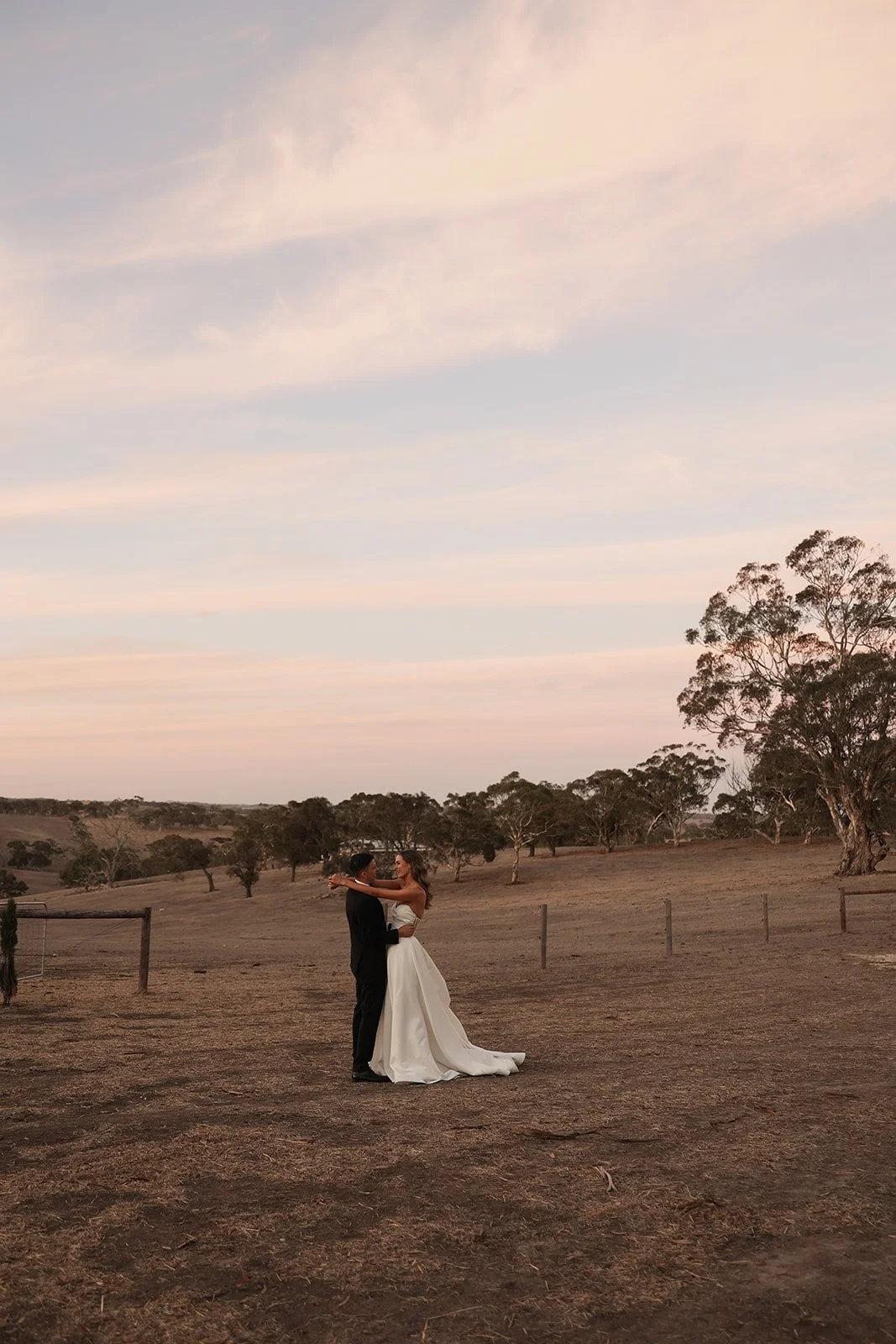 A bride and groom dancing outdoors on a dirt field at sunset, with trees and a cloudy sky in the background.