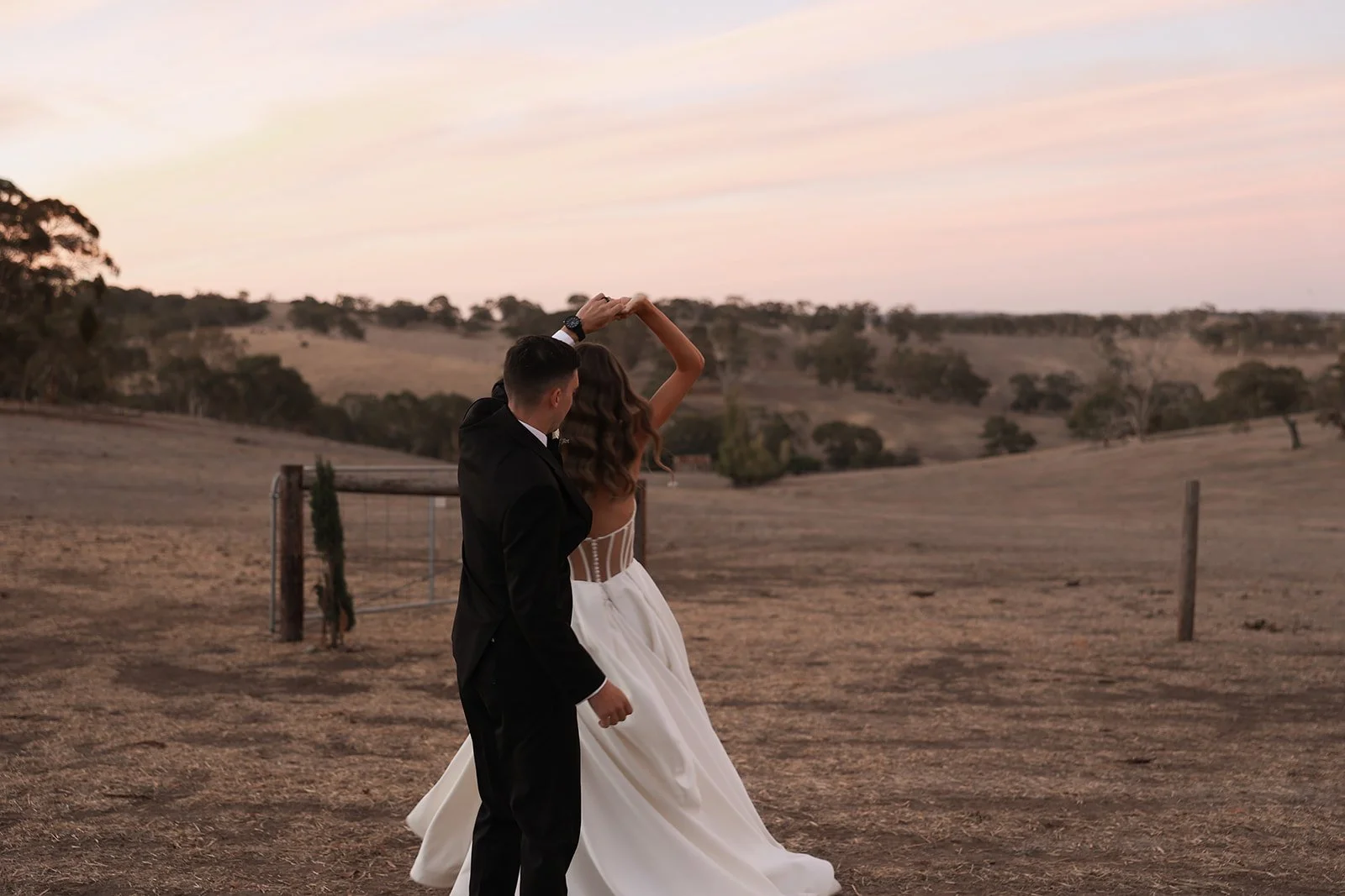 A couple dancing in a rural landscape at sunset, with rolling hills and sparse trees in the background.