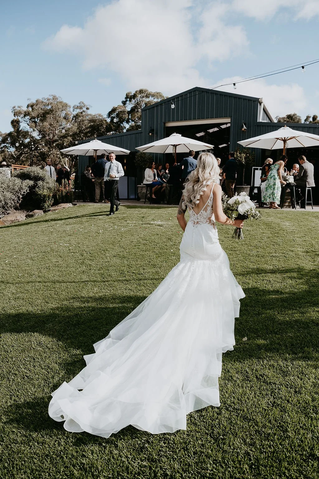A bride in a wedding gown holding a bouquet walking on grass towards an outdoor gathering with guests and blue sky overhead.