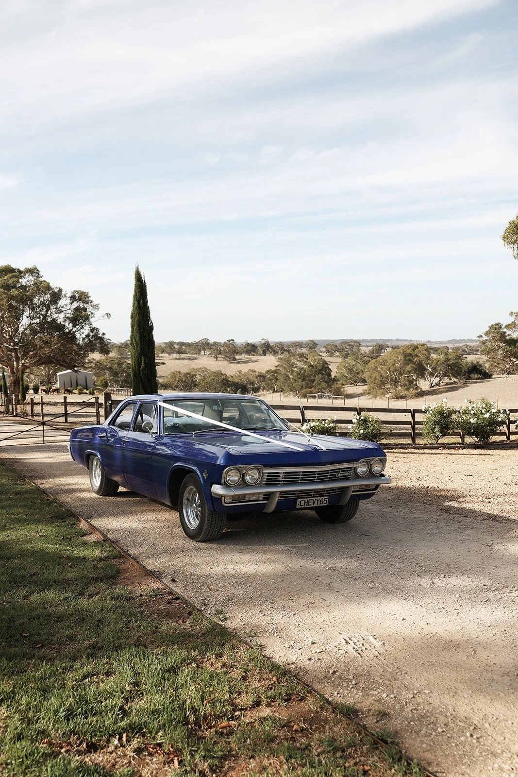 A vintage blue Chevrolet car parked on a gravel driveway in a rural landscape with trees, a fence, and open fields under a partly cloudy sky.