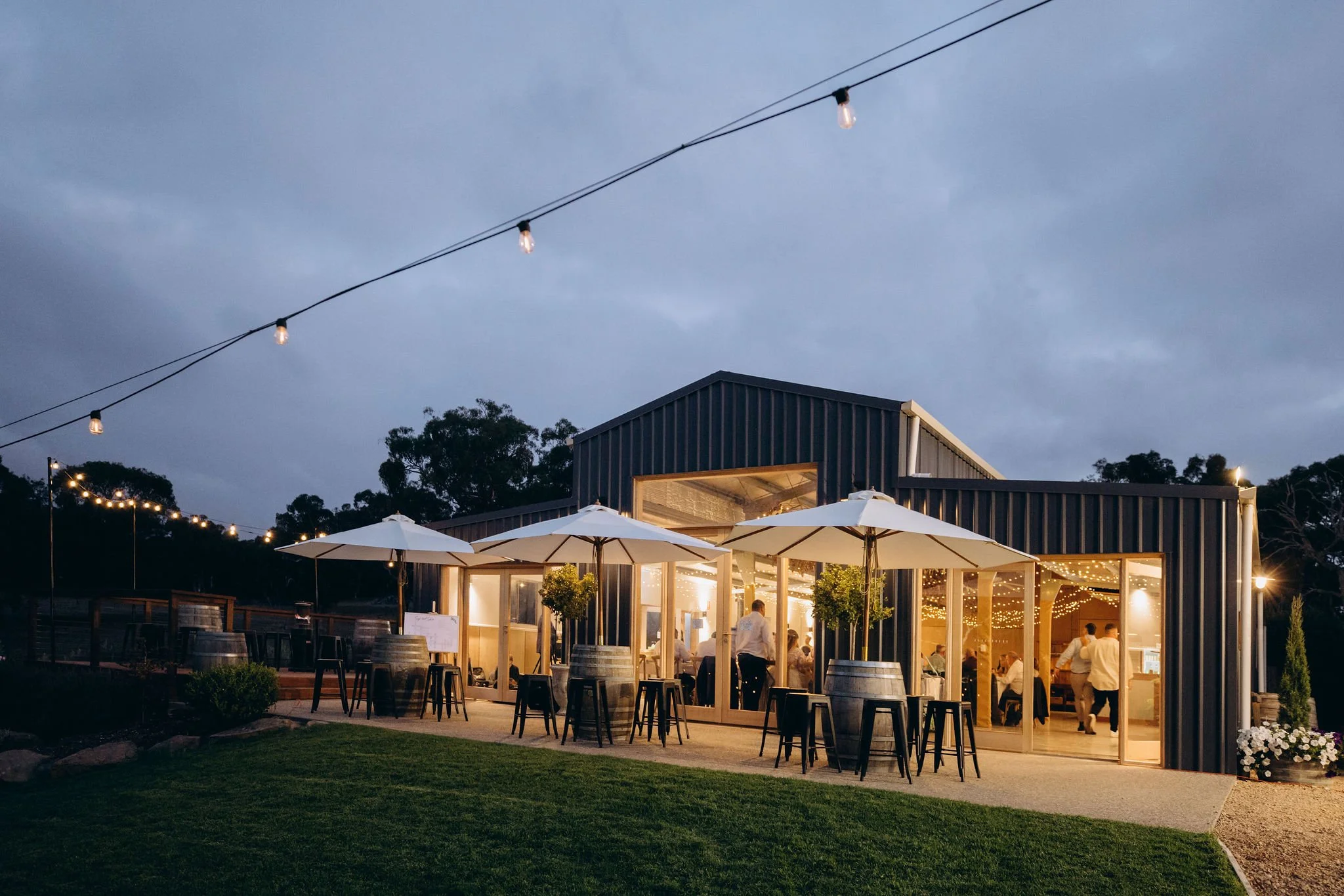 A modern event space or restaurant with large windows and an outdoor patio area, decorated with string lights and umbrellas, during evening hours.