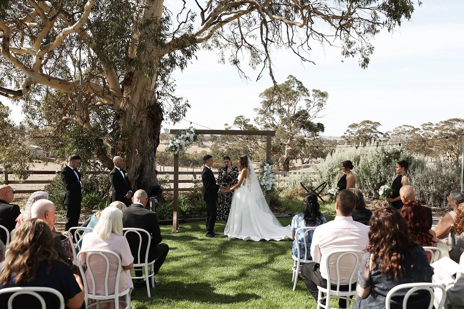 Outdoor wedding ceremony with bride and groom exchanging vows under a wooden arch decorated with white flowers, surrounded by guests seated on white chairs, in a rural setting with tall trees and blue sky.