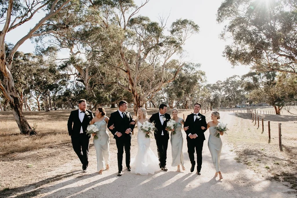 A bride and groom walking with their wedding party outdoors in a wooded area, all dressed in formal attire with the bride in a white gown and others in black suits and light-colored dresses, holding flowers.
