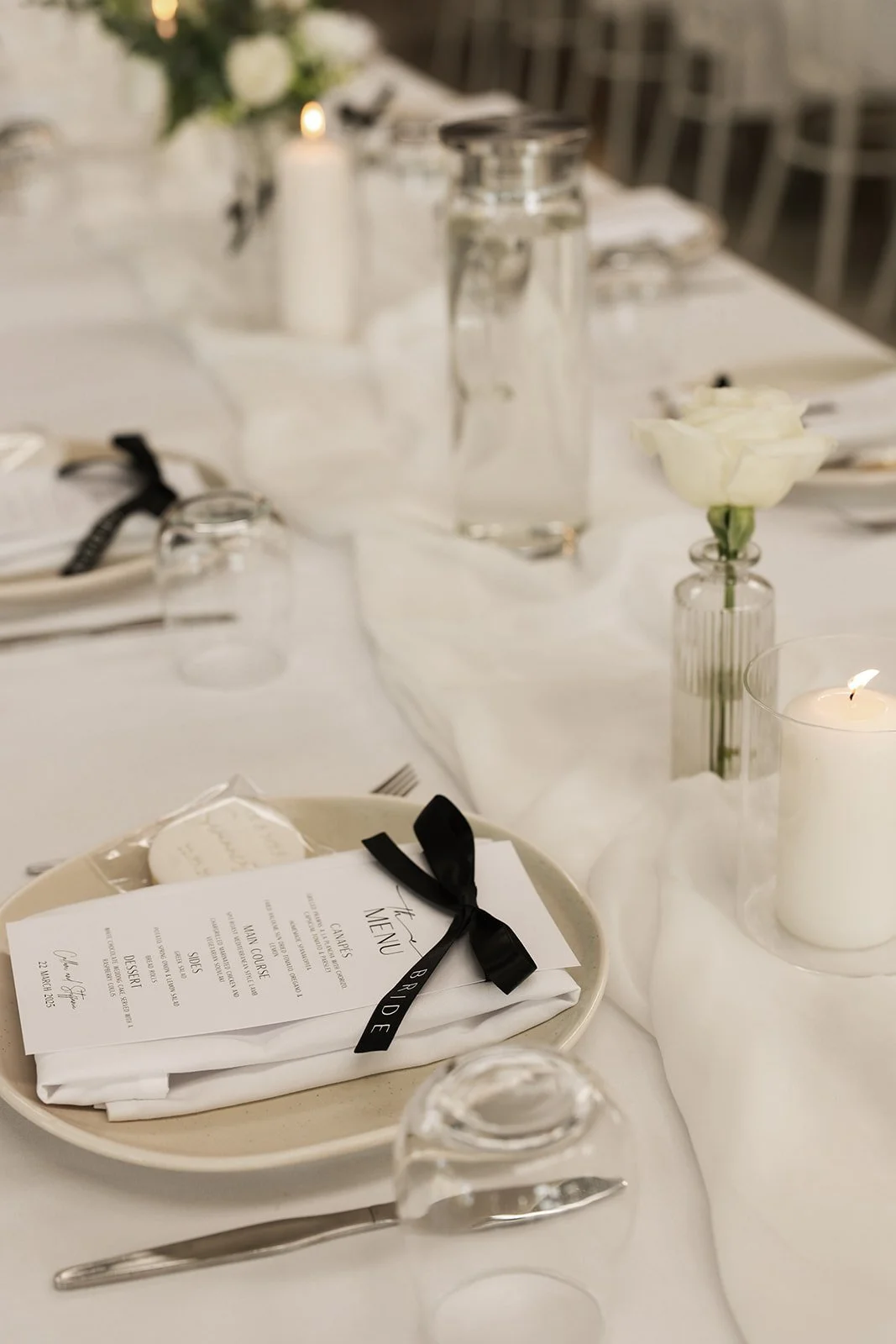 Elegant wedding reception table setting with white tablecloth, black-ribboned menu, white flower in a small glass vase, candles, and glassware.