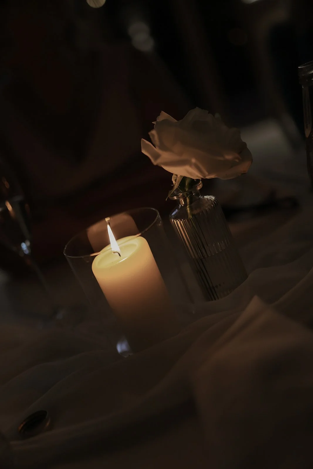 A lit candle, a white flower in a glass vase, and a piece of fabric in a dimly lit setting.