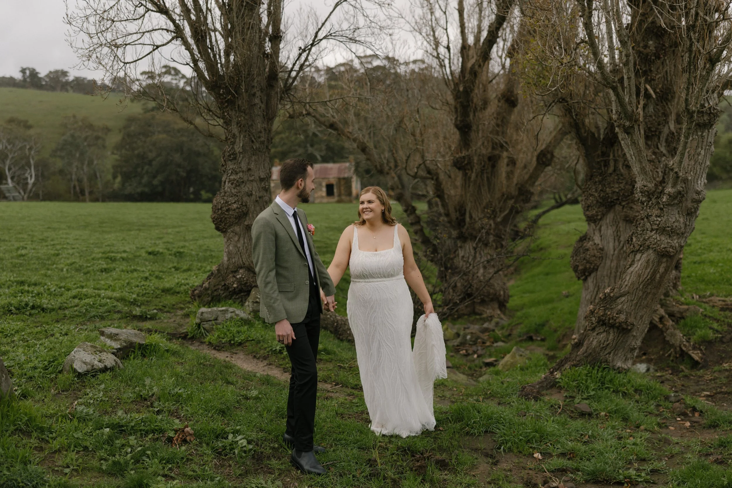 A couple holding hands and walking outdoors in a grassy field with large trees and a small building in the background, dressed in wedding attire.