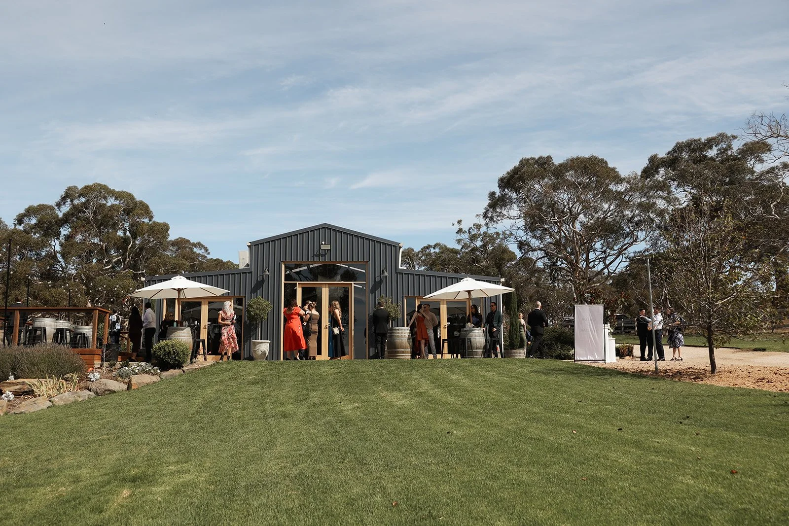 Guests socializing outside a black metal building with white umbrellas, barrels, and potted plants on a sunny day.
