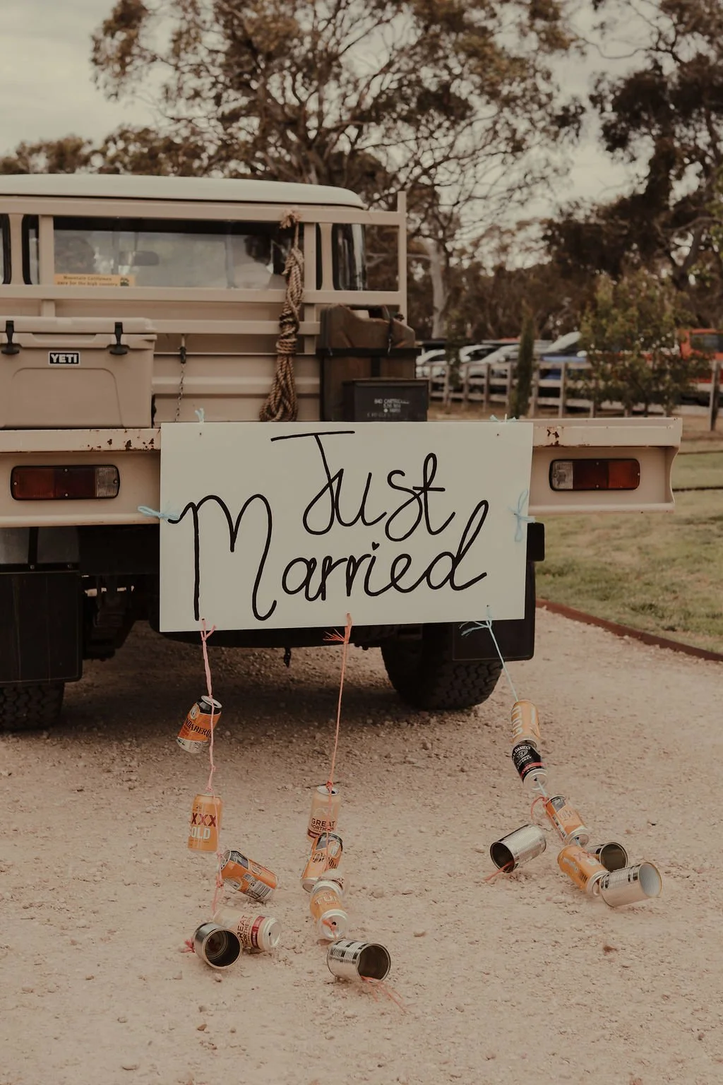 A pickup truck with a large sign that reads 'just married' attached to the back, with empty cans hanging from pink strings. The truck is parked outdoors on a dirt surface with trees and parked cars in the background.