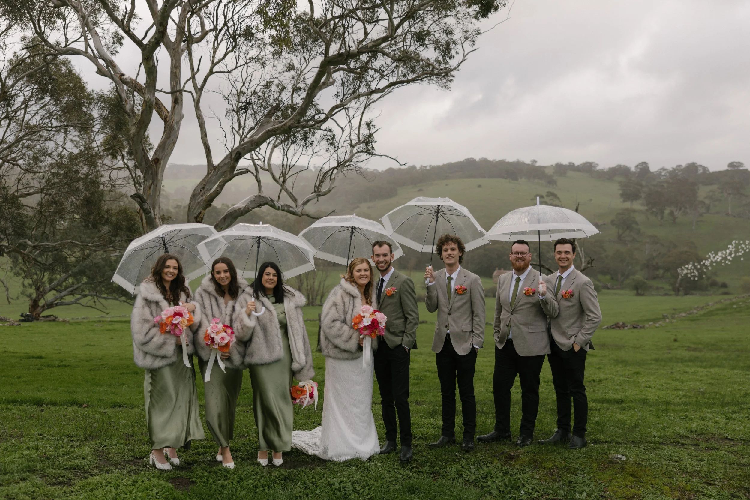 A wedding party standing outdoors in a green field on a rainy day, holding transparent umbrellas. The group includes women in matching green dresses with white fur shawls, and men in gray suits with boutonnières. The bride is holding a bouquet, and t