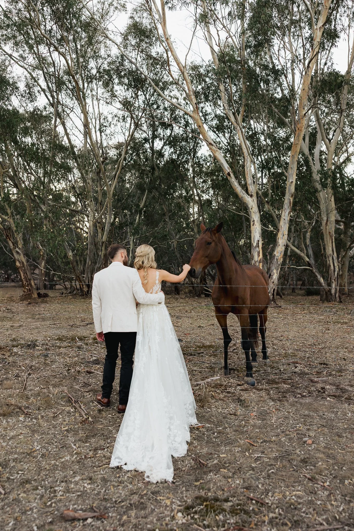 A bride and groom in wedding attire standing near a brown horse in a wooded outdoor setting.