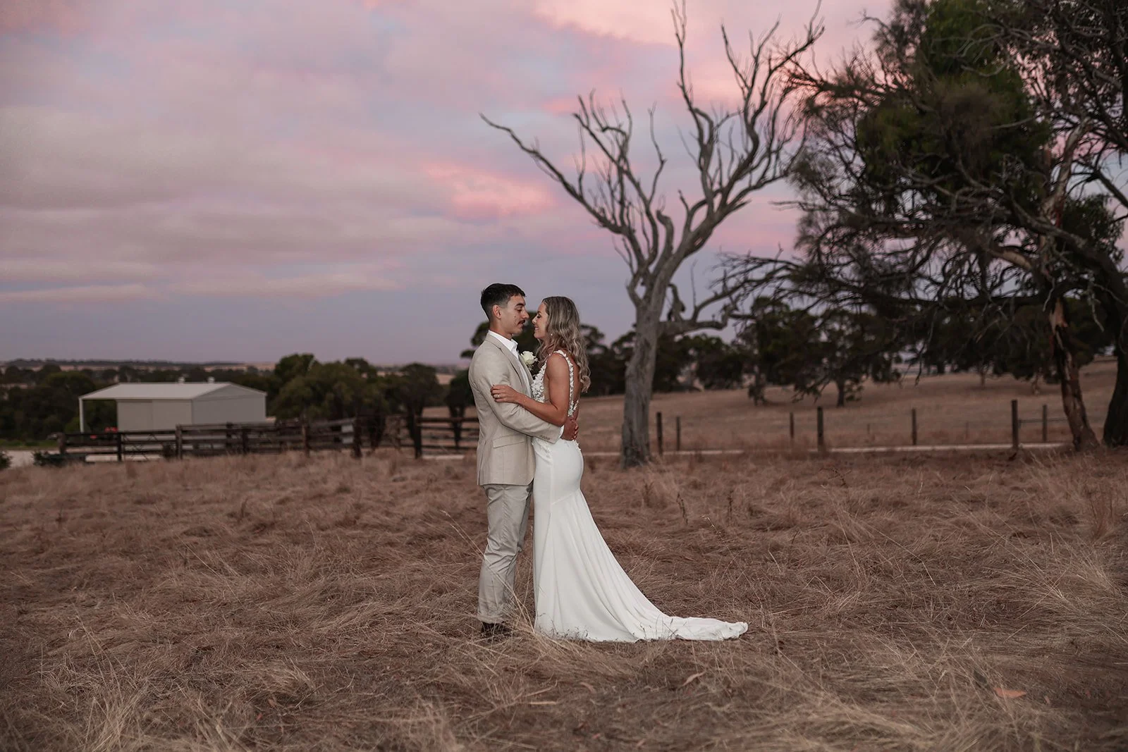 A bride and groom in wedding attire embrace in a field at sunset, with trees and a fence in the background.