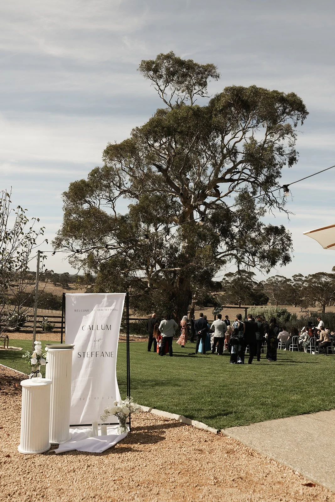 Wedding guests gather outdoors near a large tree on a grassy area, with a white sign welcoming Callum and Steffanie, decorated with flowers, in a rural setting with a partly cloudy sky.