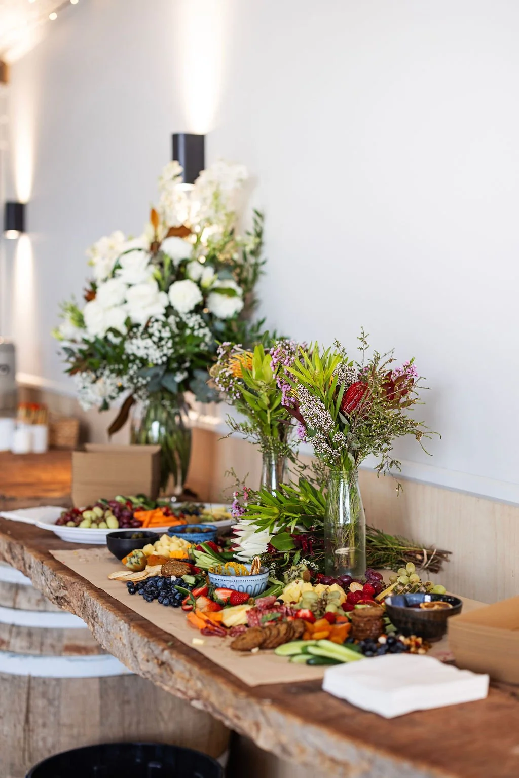 A rustic wooden table with a fruit and snack spread, decorated with floral arrangements in glass vases, set against a plain white wall.