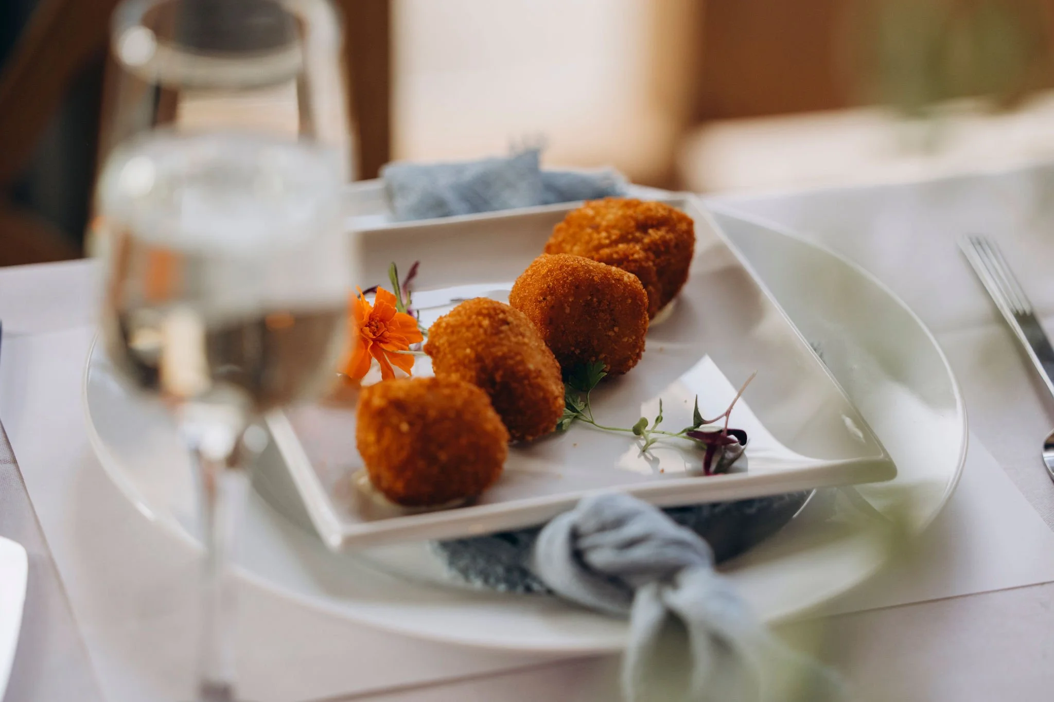 Plate with four golden-brown fried appetizers garnished with small orange and purple edible flowers, on a white tablecloth with a glass of water in the foreground.