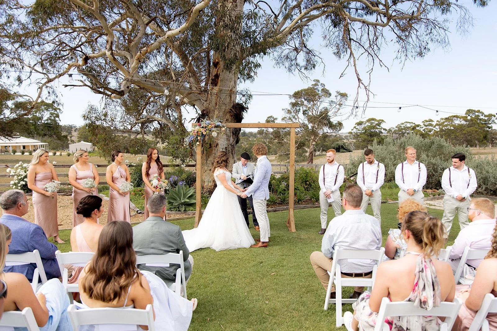 Outdoor wedding ceremony with bride and groom exchanging vows under a wooden arch decorated with flowers, surrounded by bridesmaids in pink dresses and groomsmen in white shirts and suspenders, seated guests on white chairs, large tree and scenic rur