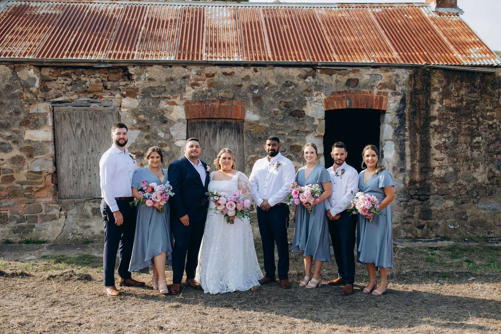 Wedding party standing in front of an old stone building with a rusty metal roof, including the bride, groom, and six bridesmaids and groomsmen holding bouquets and dressed in formal attire.
