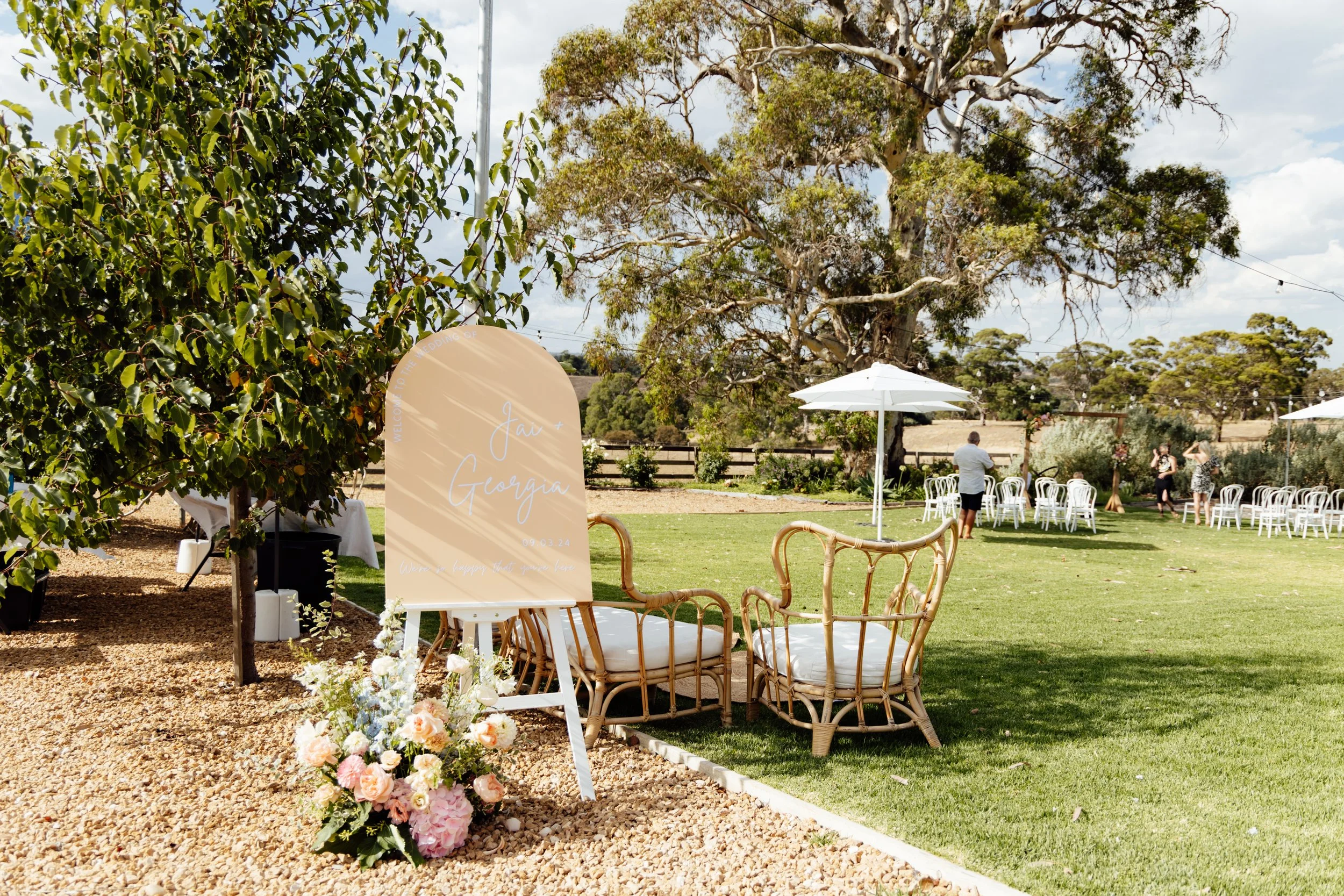 Outdoor wedding setup with white chairs arranged in the background, a white umbrella, a brown wicker loveseat and chair with white cushions, a floral arrangement with pink and white flowers, and a large tree in a garden setting.