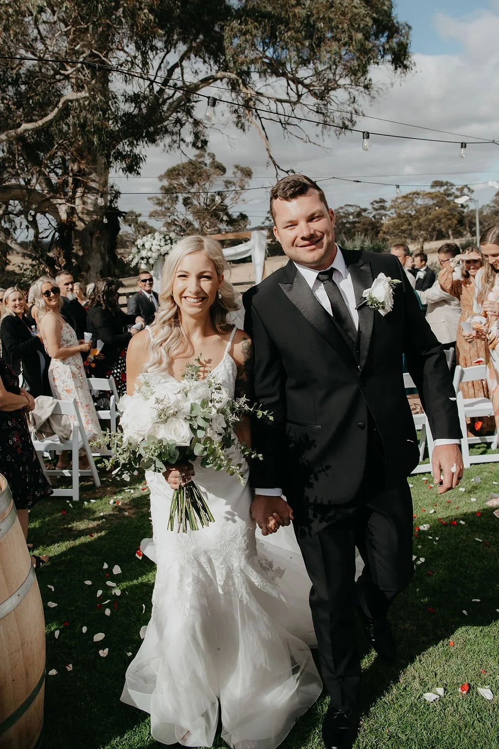 A newlywed couple walking hand-in-hand outdoors at their wedding ceremony, surrounded by guests and decorated with string lights and floral arrangements, on a sunny day with a partly cloudy sky.