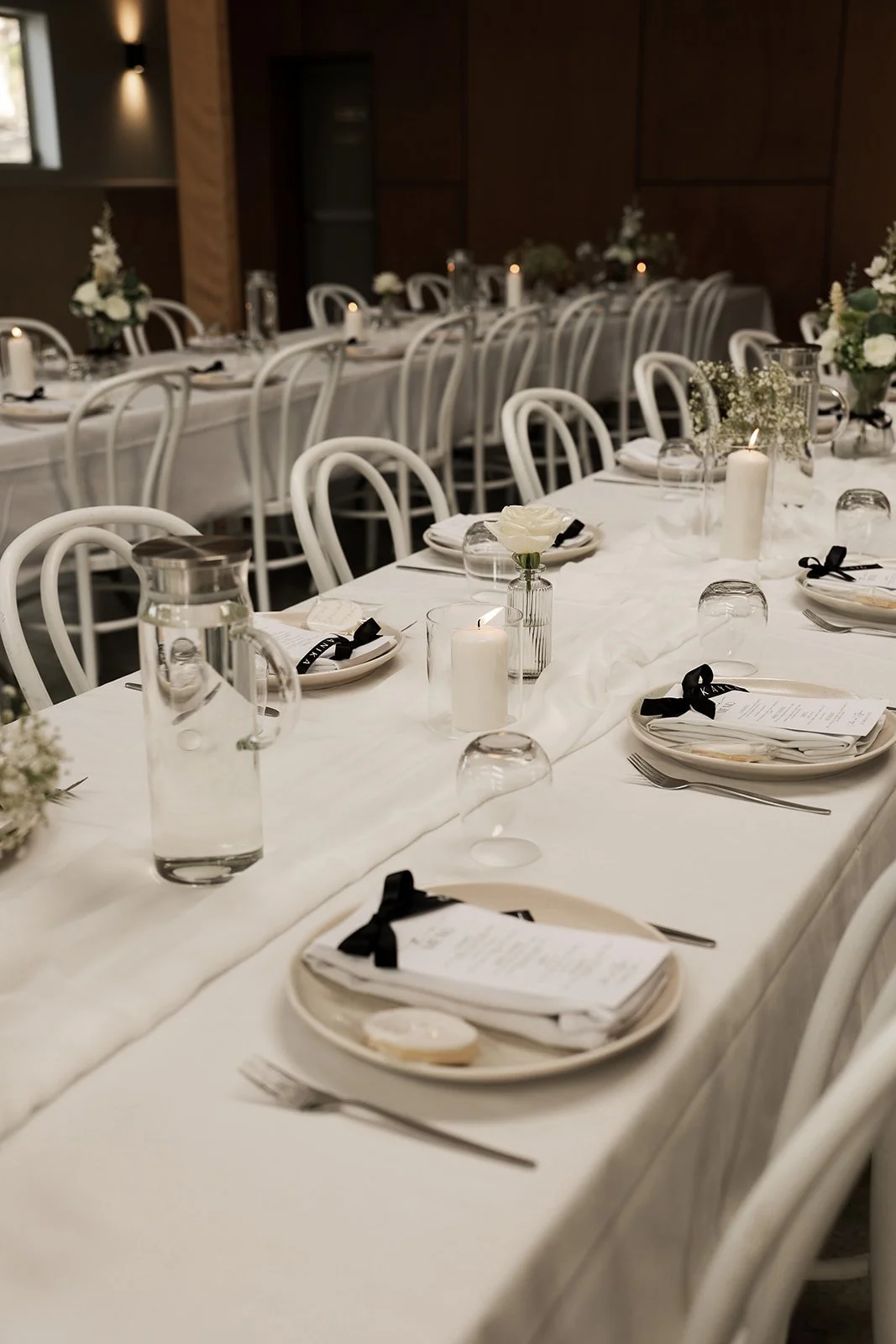 Elegant banquet table set with white tablecloth, white plates with black ribbons, silverware, glass water pitchers, candles, and floral centerpieces in a decorated room.