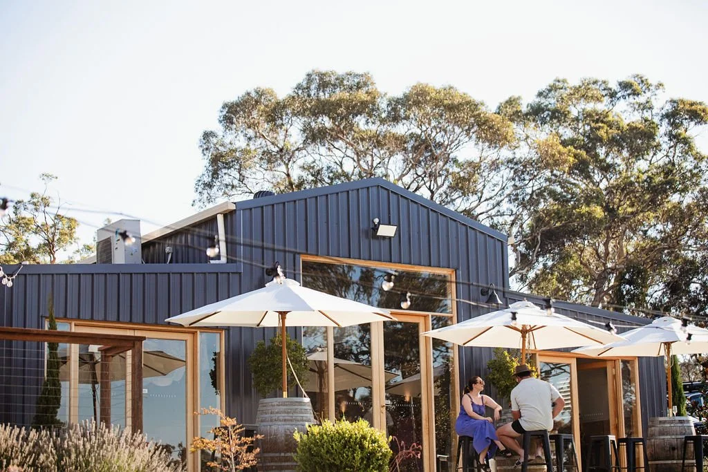 People sitting outdoors at a restaurant or bar with white umbrellas, in front of a modern dark blue building with large windows, surrounded by trees.