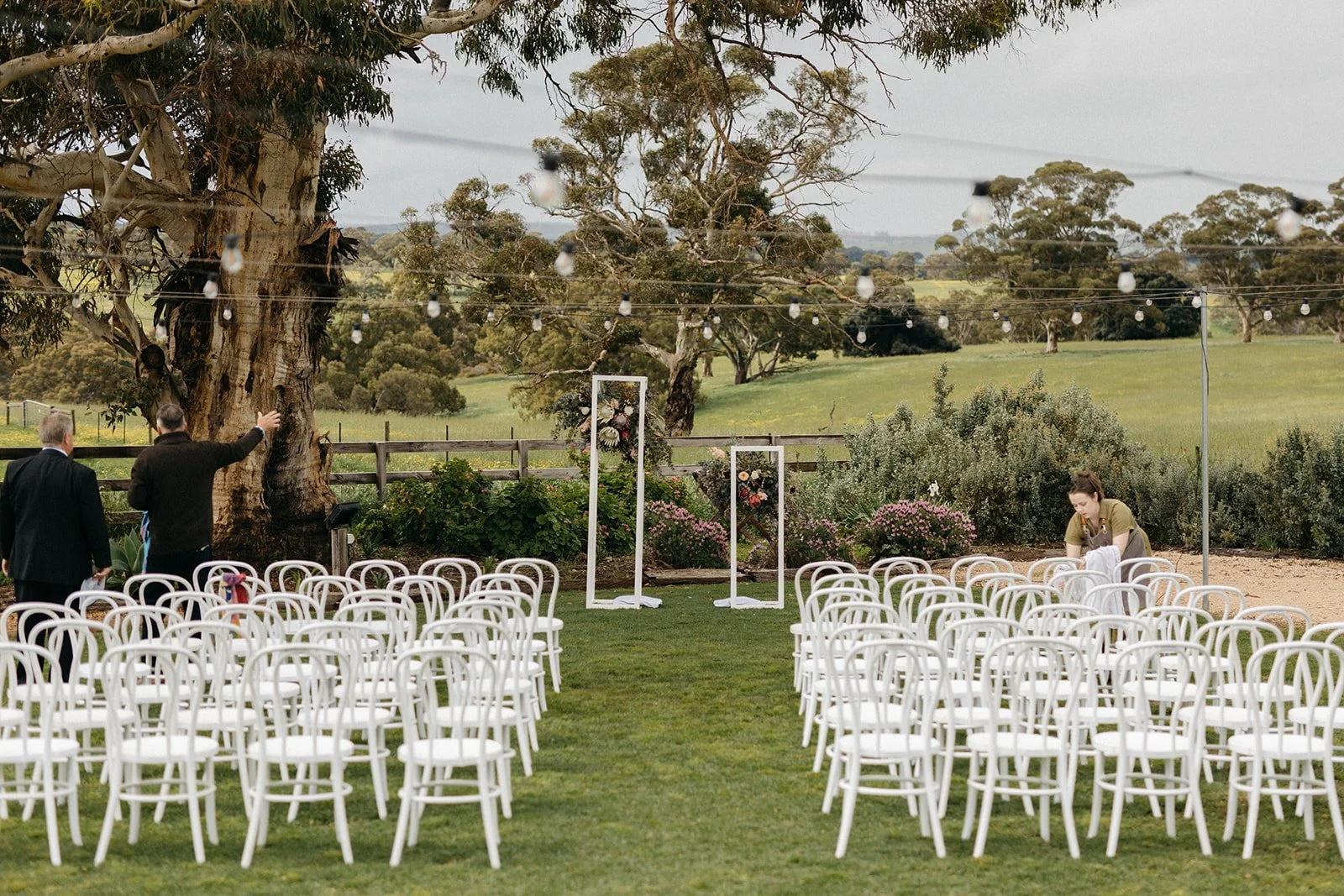 Outdoor wedding setup with white chairs arranged in rows on green grass, floral decorations, and a large tree in a rural landscape. There are few people preparing for the event, with colorful string lights hanging overhead.