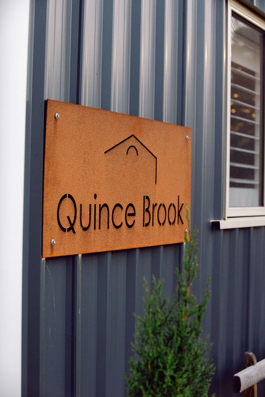 Rust-colored sign with black text reading "Quince Brook" and a simple line drawing of a house, mounted on a gray metal wall with a window nearby.