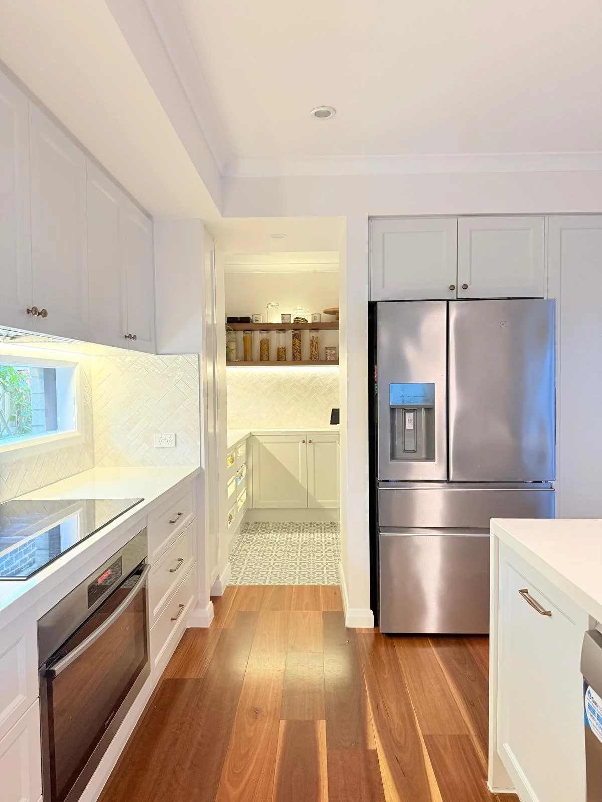 Modern kitchen with white cabinets, stainless steel refrigerator, wood flooring, and a small window above the counter, leading to a pantry area with open shelves and a tiled floor.