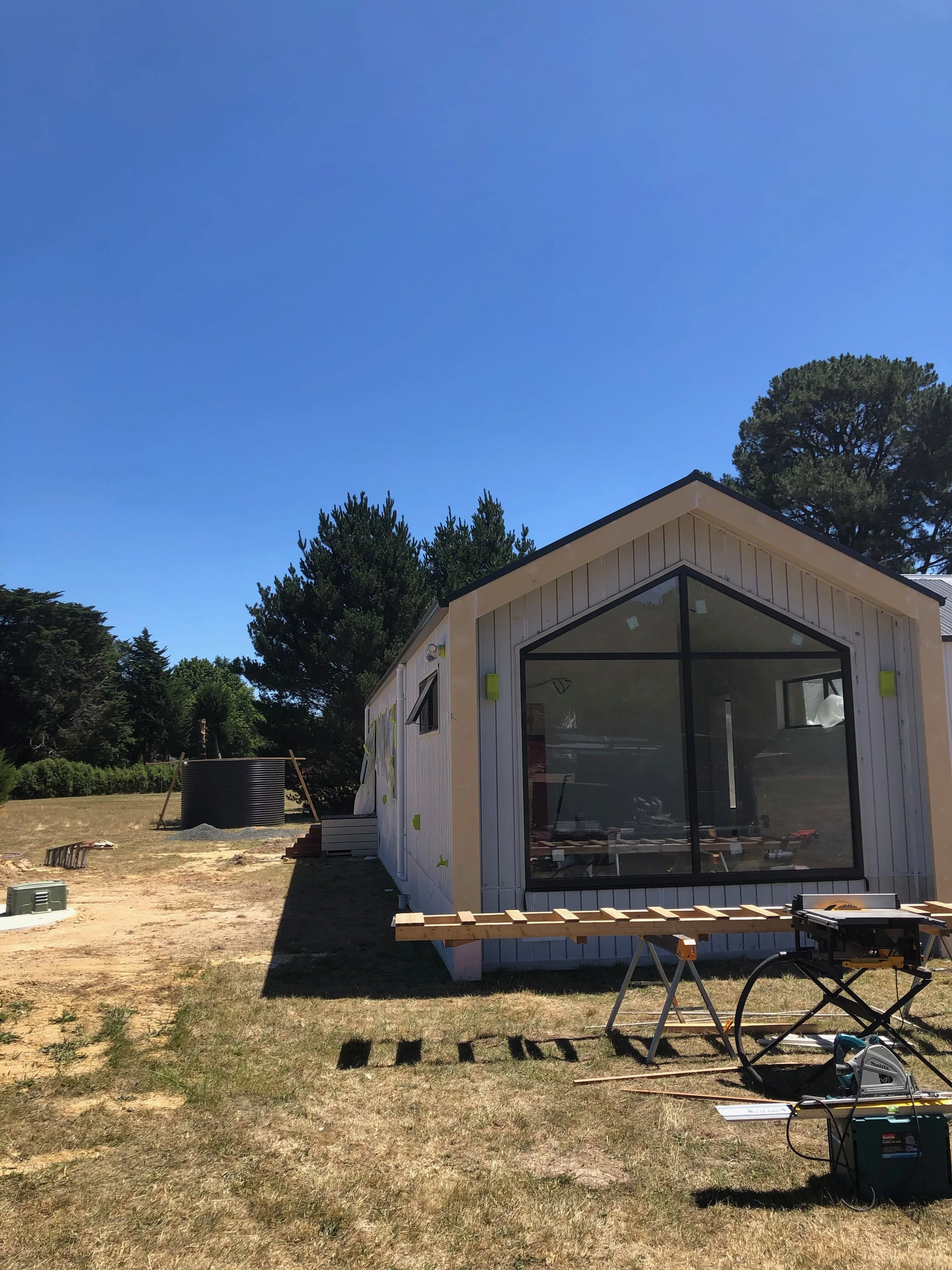 Under construction tiny house on a grassy field with tools and a water tank, trees in the background, clear blue sky.