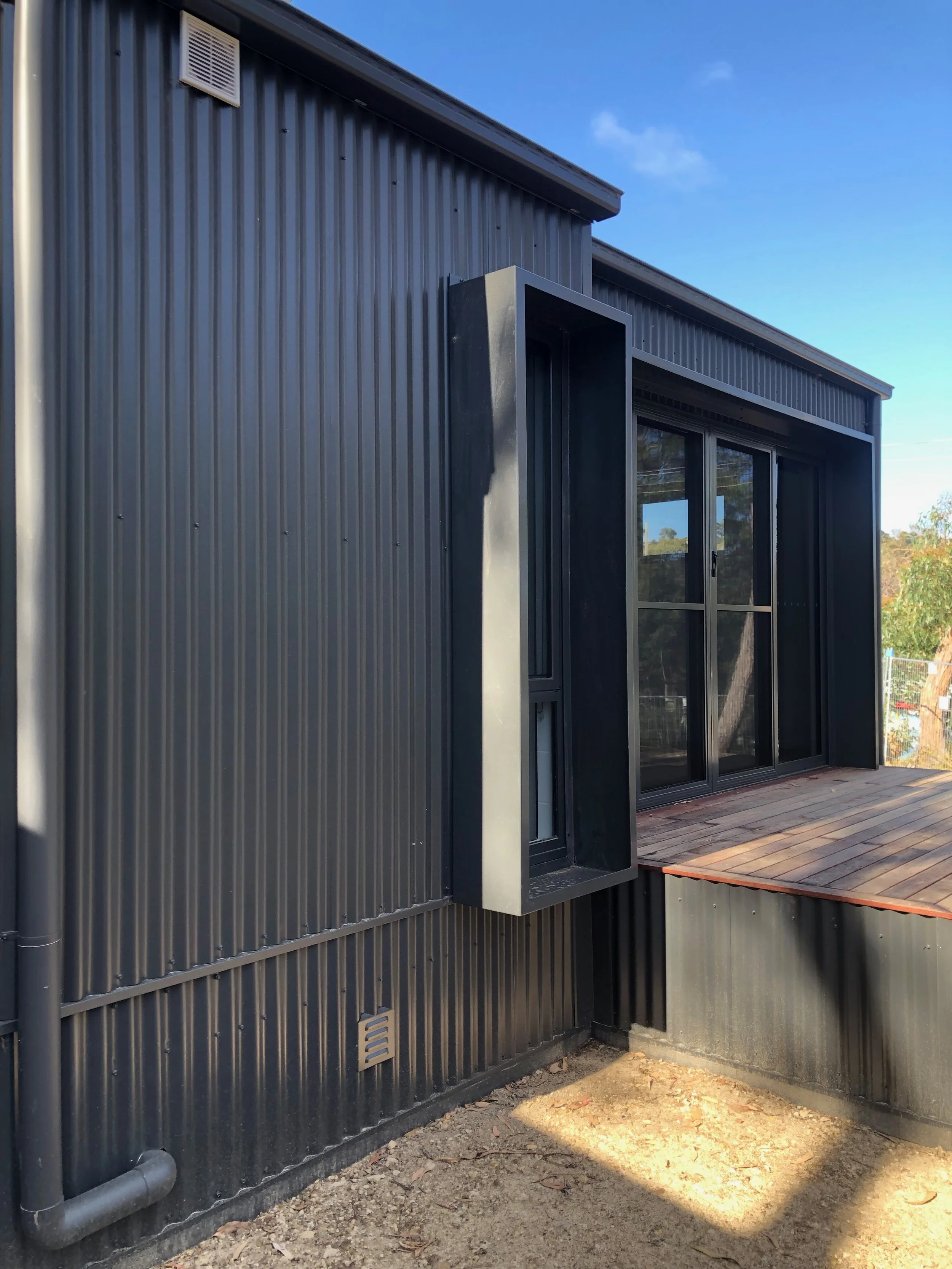 Exterior of a modern building with black metal siding, large sliding glass doors, a small wooden deck, and an angular black awning above the window.