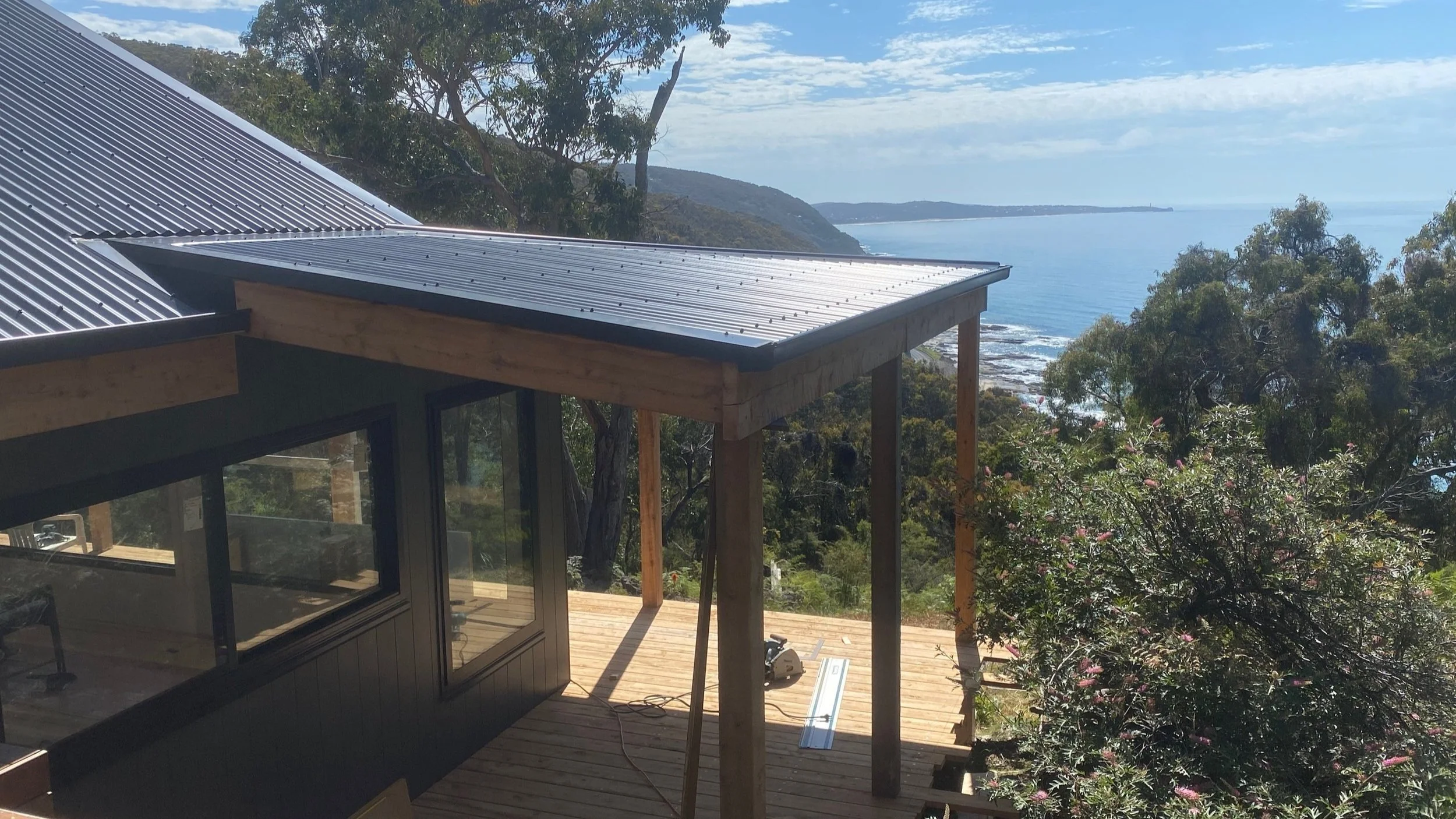 Partially constructed house with a metal roof, large windows, and a wooden deck overlooking the ocean and trees.