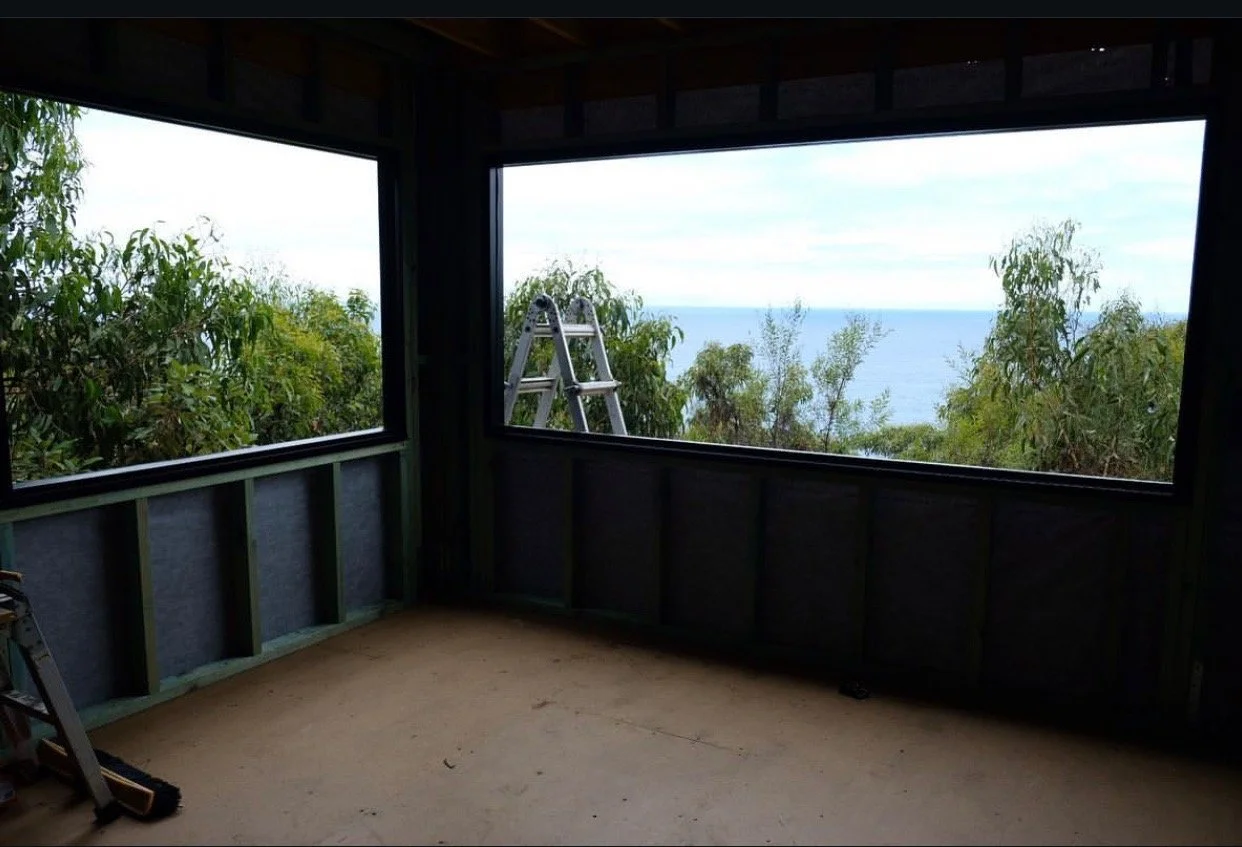 View from an unfinished room with large open windows showing trees and a distant ocean, with a ladder and construction materials inside.