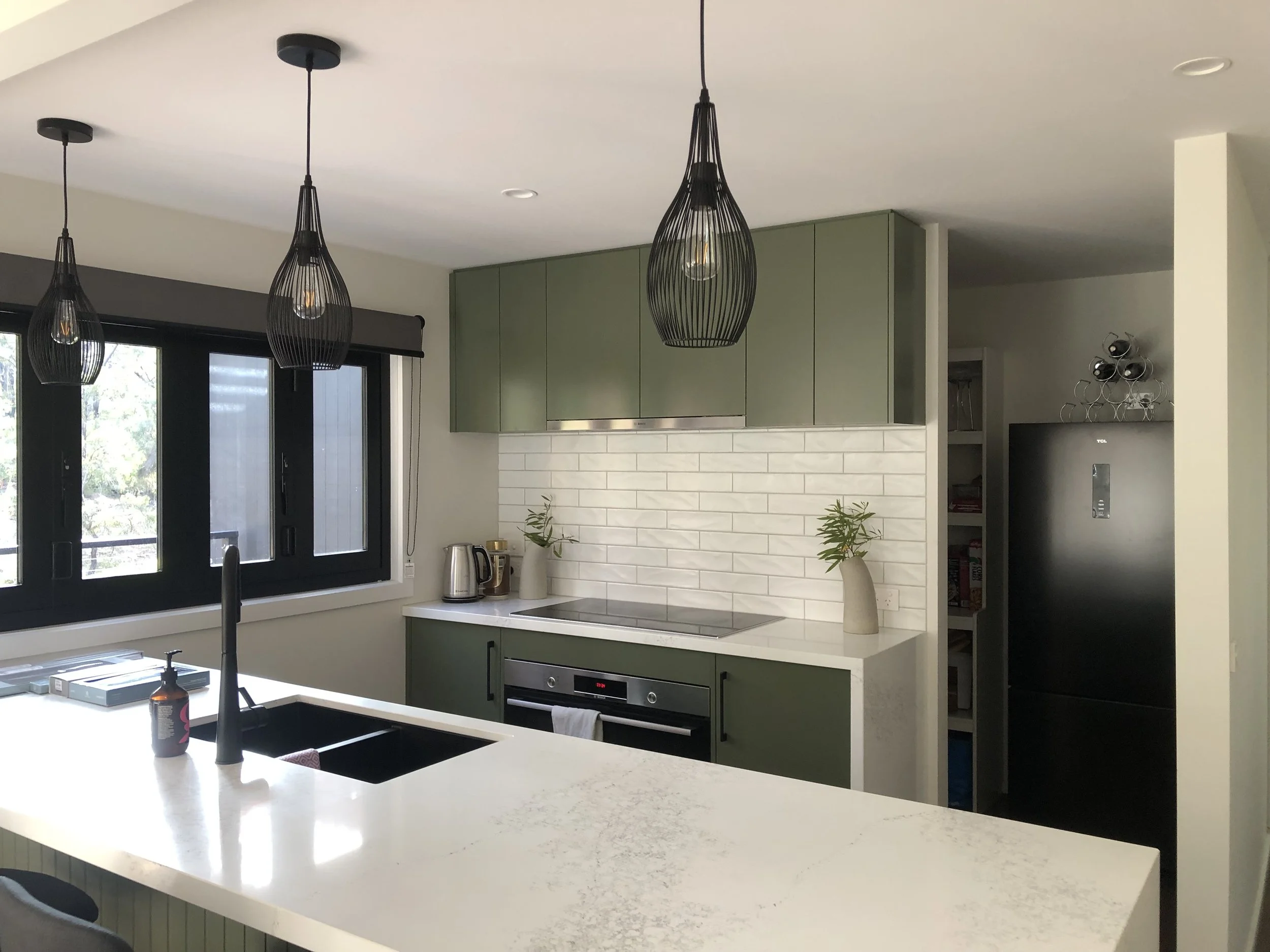Modern kitchen with green upper cabinets, white countertop, black window frames, black pendant lights, and a white brick backsplash. Contains a sink, electric stove, toaster, potted plants, and a black refrigerator.