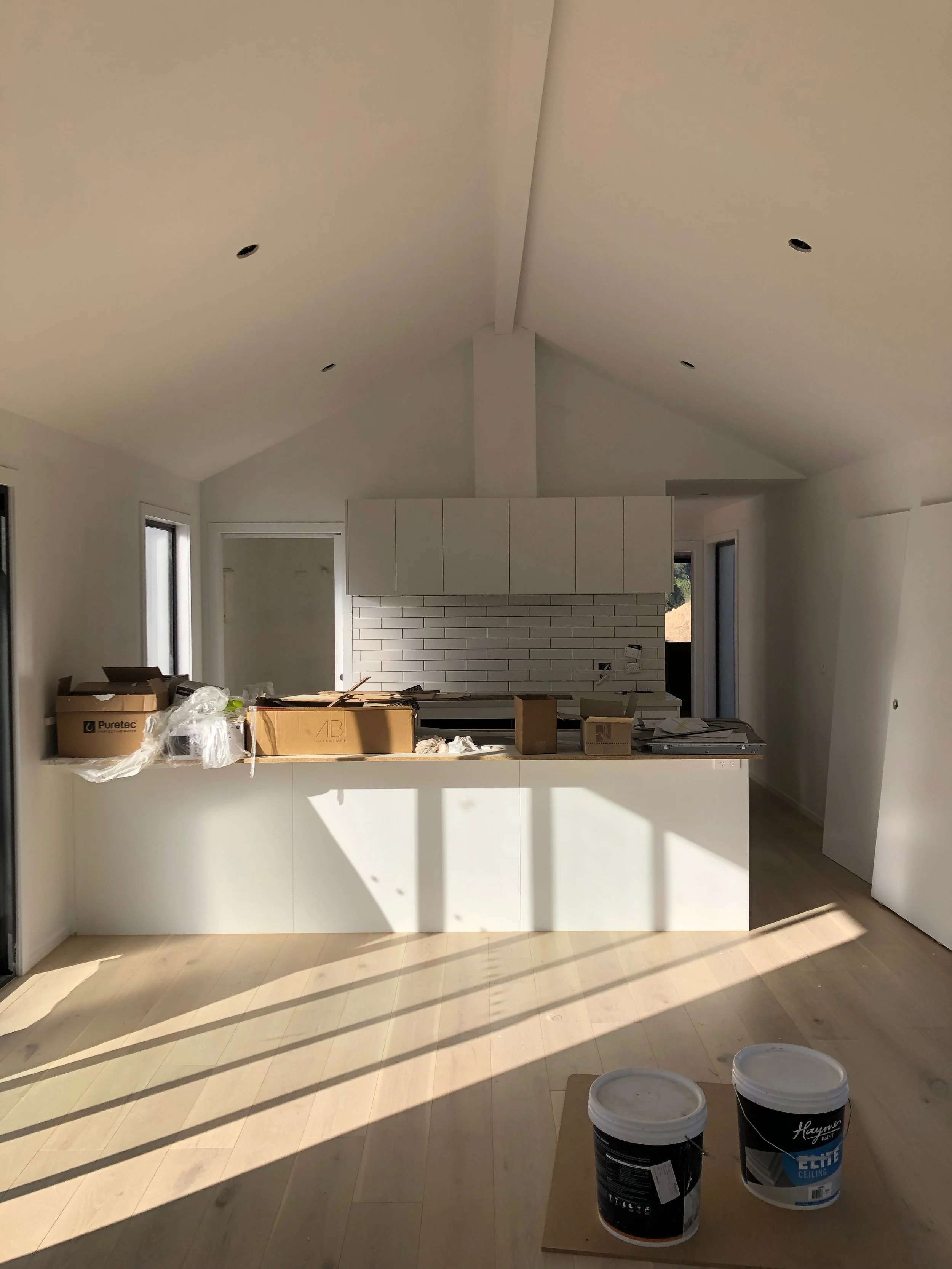 Empty kitchen under construction with unopened paint buckets on a small table in front, natural light coming through large windows, white cabinets and tiles, and construction materials on the counter.