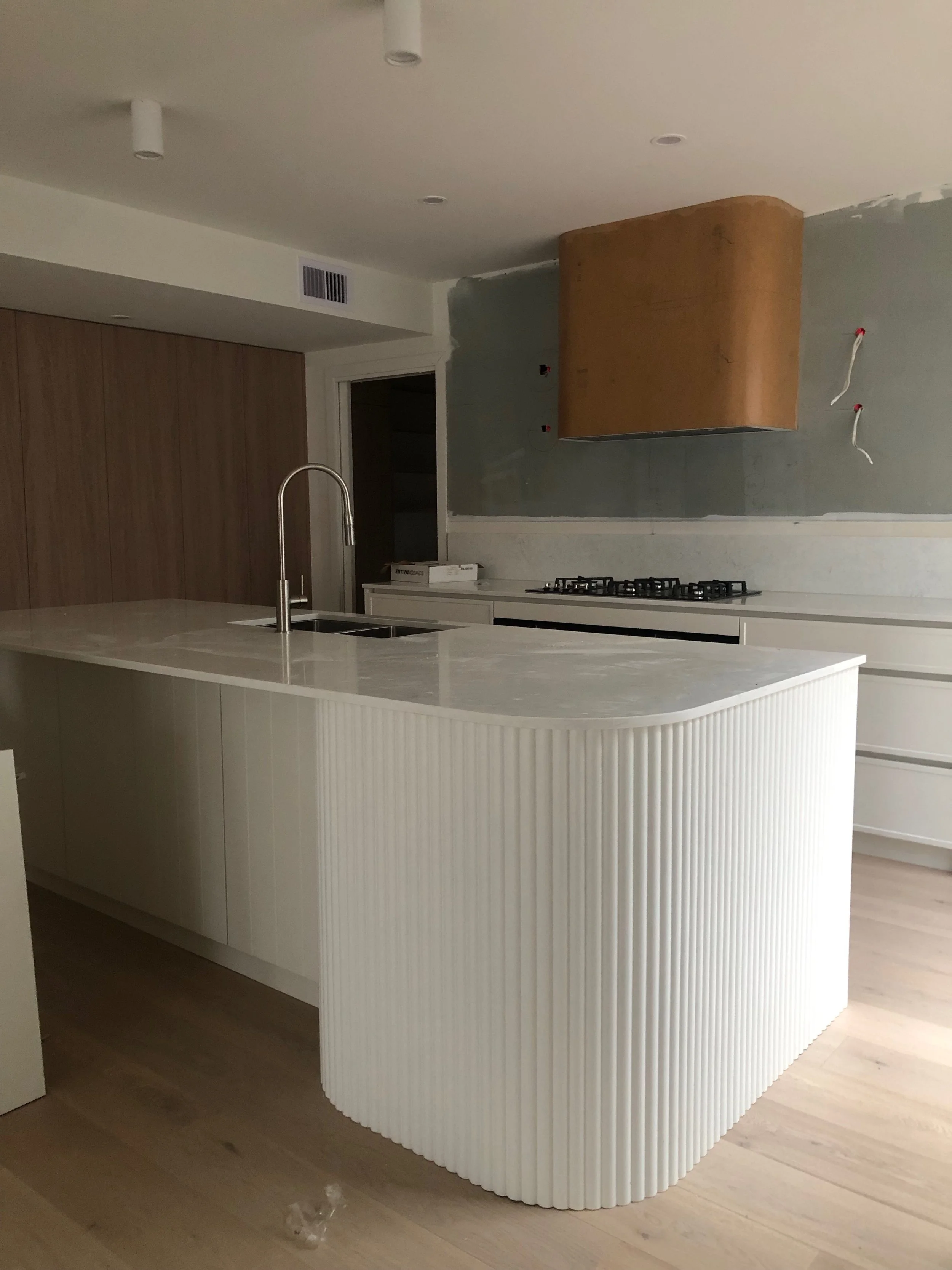 Modern kitchen with white curved island, stainless steel faucet, wooden cabinet range hood, and a gray wall with electrical wires.