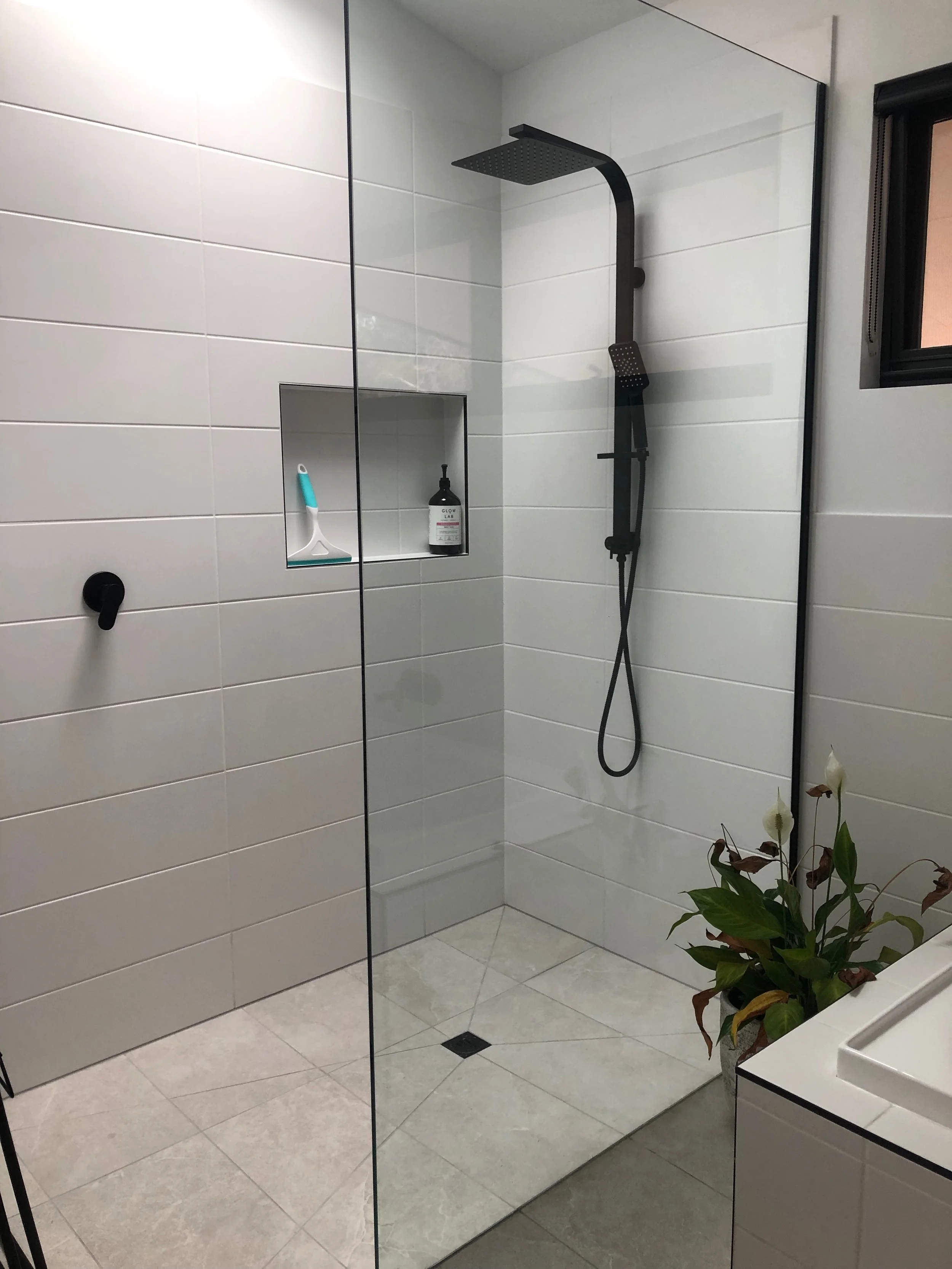 Modern shower with black rain showerhead and handheld shower in a tiled bathroom, with a built-in shelf holding cleaning supplies, a window, and a potted plant outside the shower.