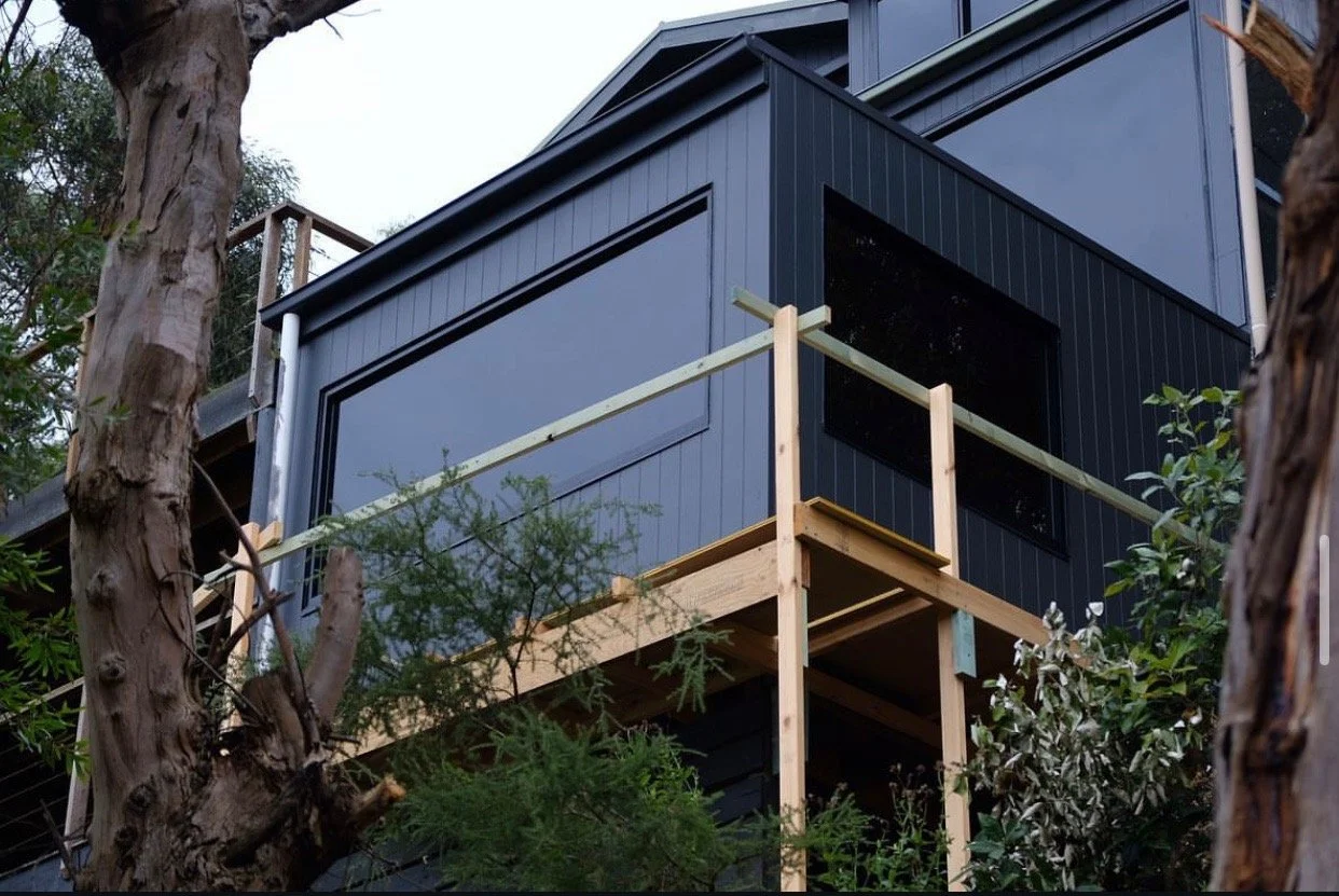 Close-up of a modern black house under construction with a wooden deck, surrounded by trees.