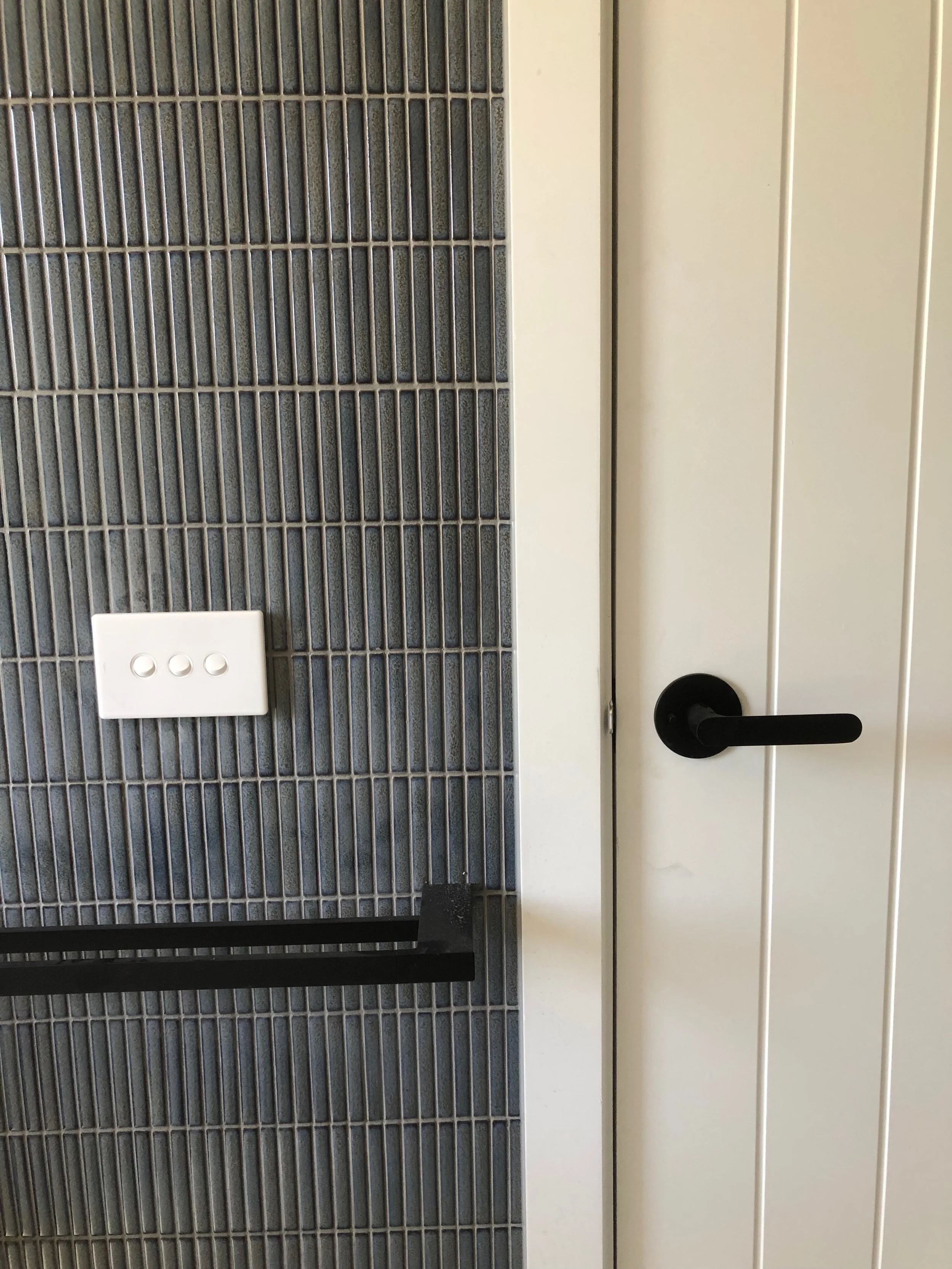 Indoor scene showing a white wall with vertical paneling, a black door handle, a section of gray tiled wall, a white three-hole switch plate, and a black horizontal bar.