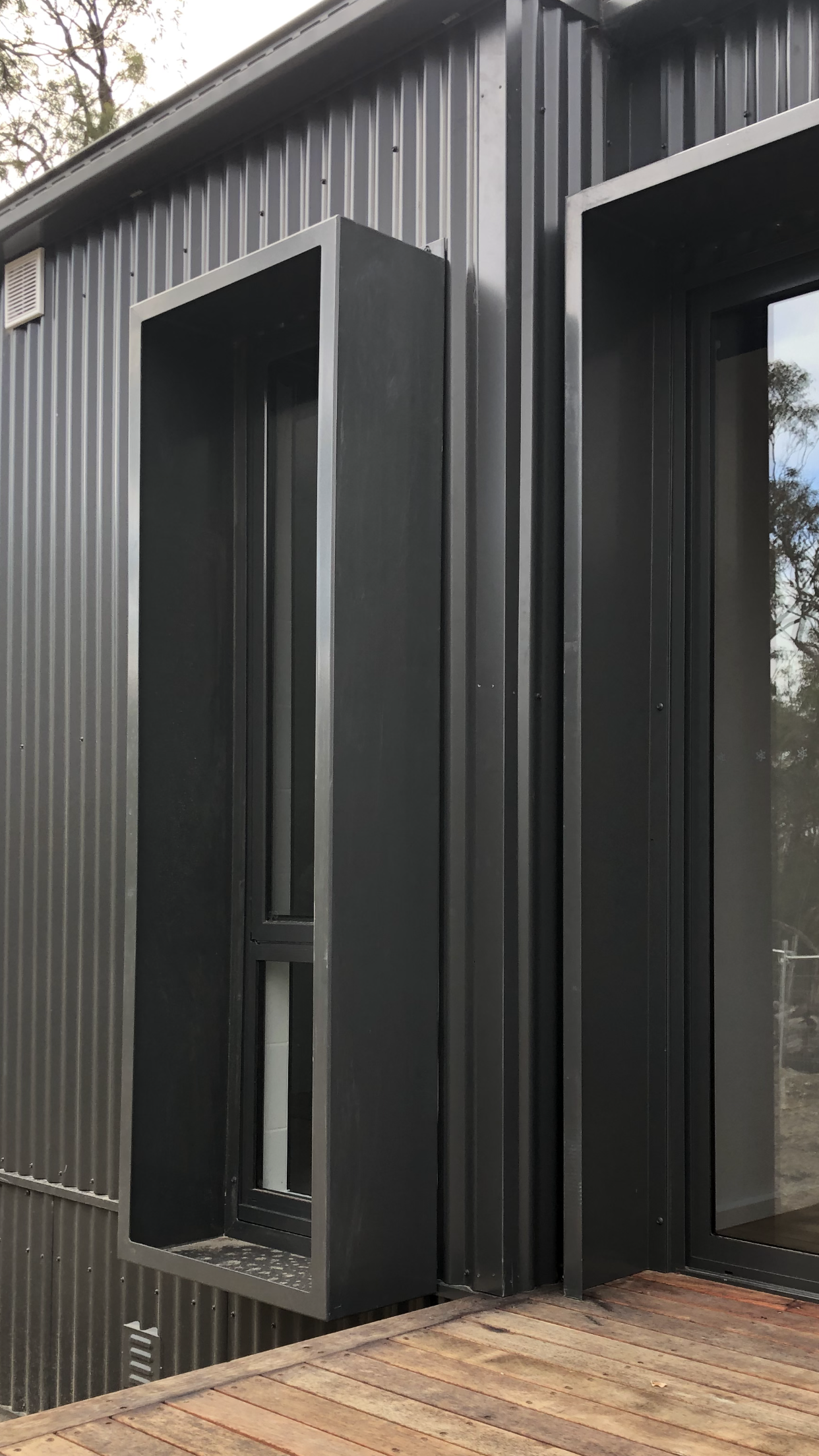 Modern building exterior with black corrugated metal siding, a narrow window with a black frame, and a black framed glass sliding door on a wooden deck.