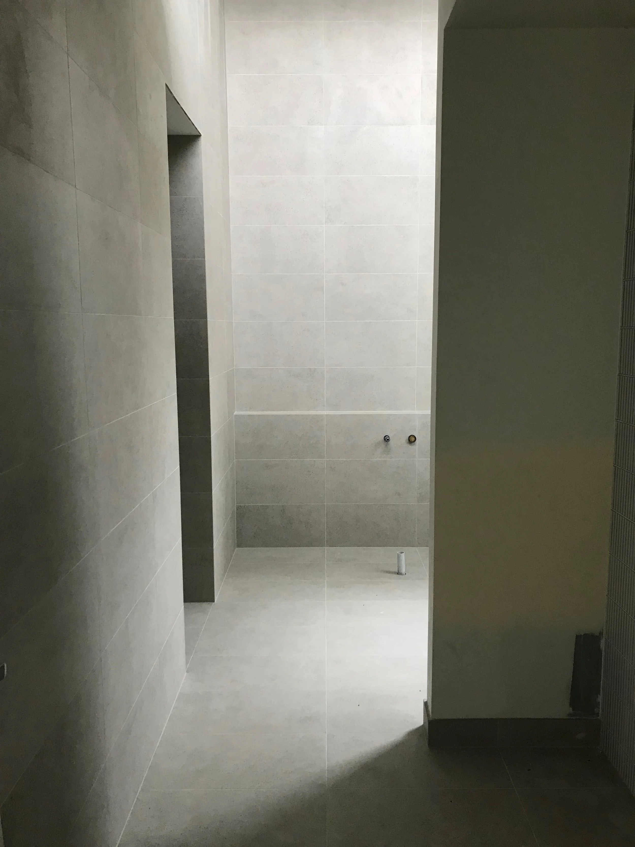 Empty bathroom with gray tiles, an open space for a shower or toilet, two water connection pipes on the wall, and a white pipe on the floor