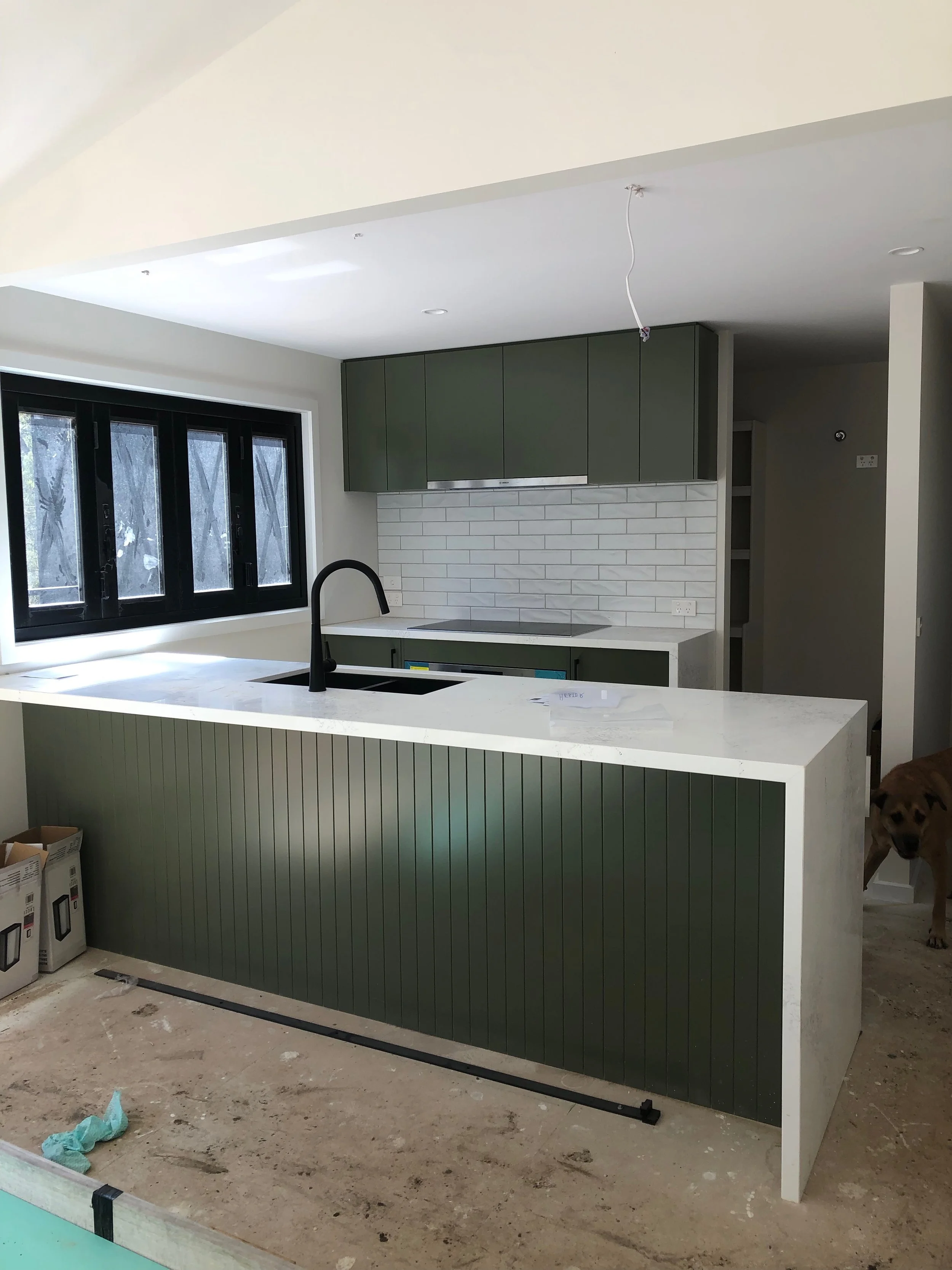 Modern kitchen under construction with green cabinetry, white subway tile backsplash, and a white countertop island with a black sink and faucet. A large window with black frames, and a dog standing in the doorway to the right.