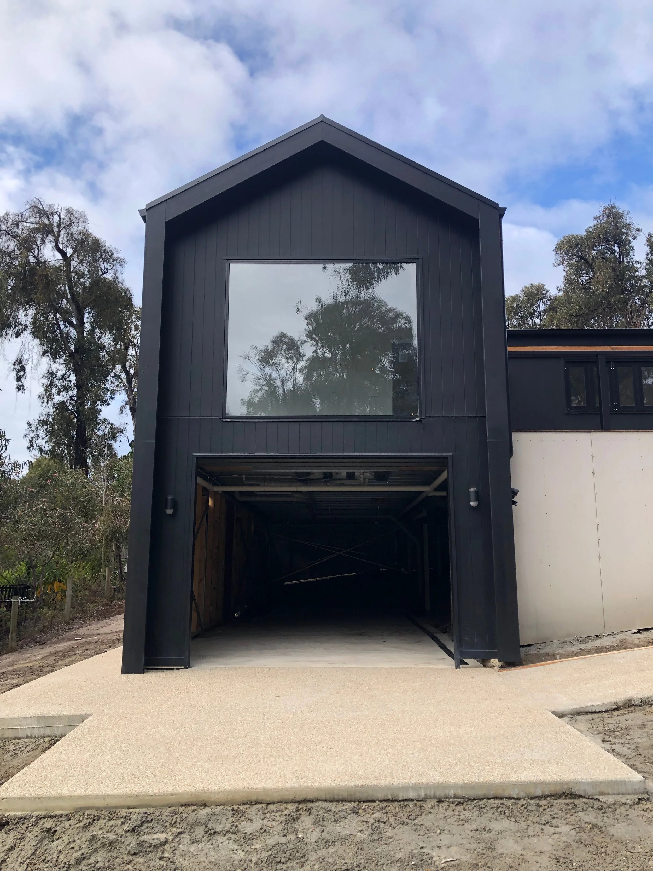 Modern black two-story house with a large garage entrance, large upper window reflecting trees, and a textured concrete driveway leading to the garage.