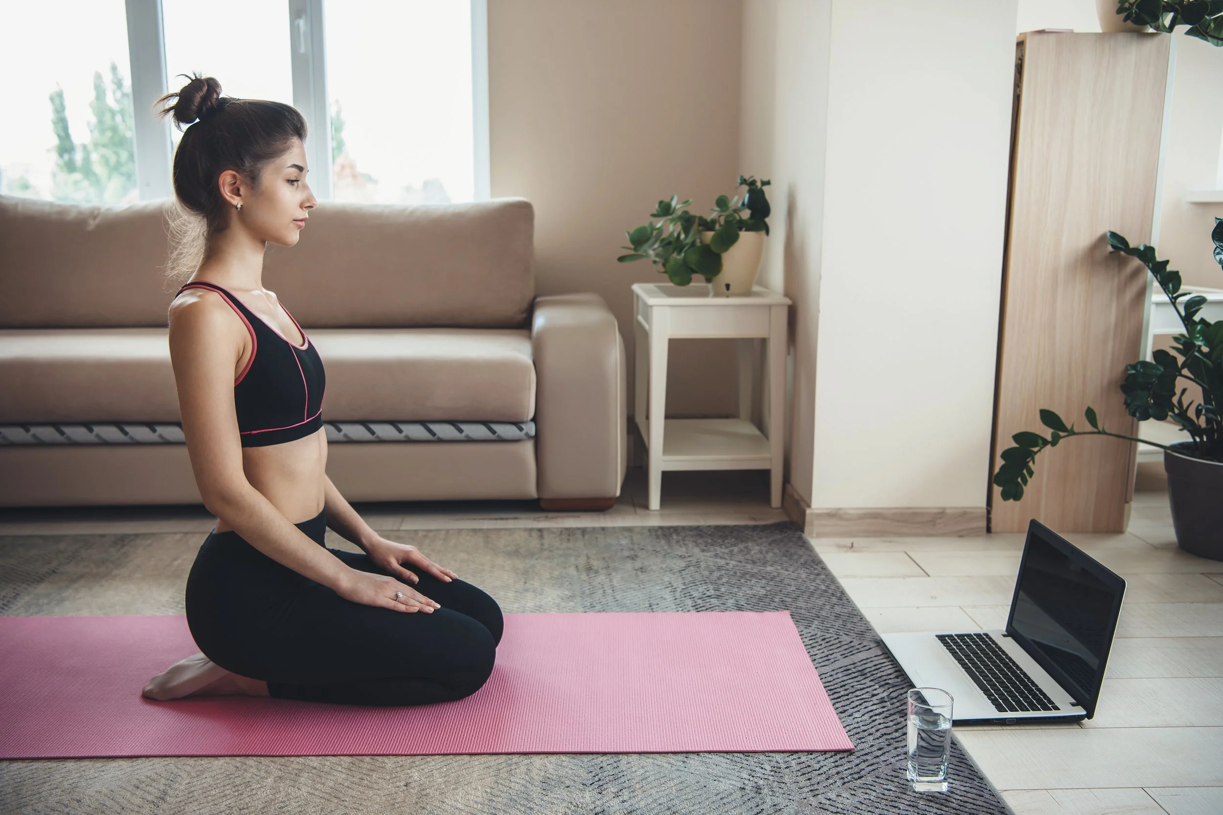 A young woman practicing yoga indoors on a pink yoga mat, sitting cross-legged with her hands on her knees, in front of a laptop and a glass of water, with a beige sofa and houseplants in the background.