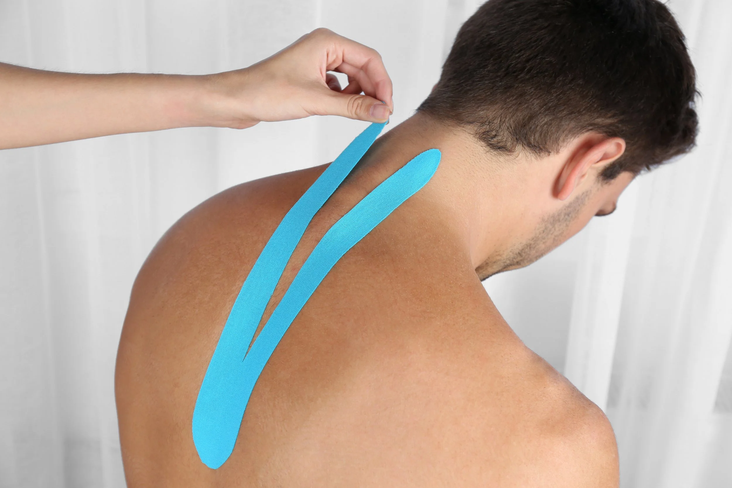 A man receiving a shoulder massage with two blue kinesiology tape strips applied to his upper back near the shoulder blade.