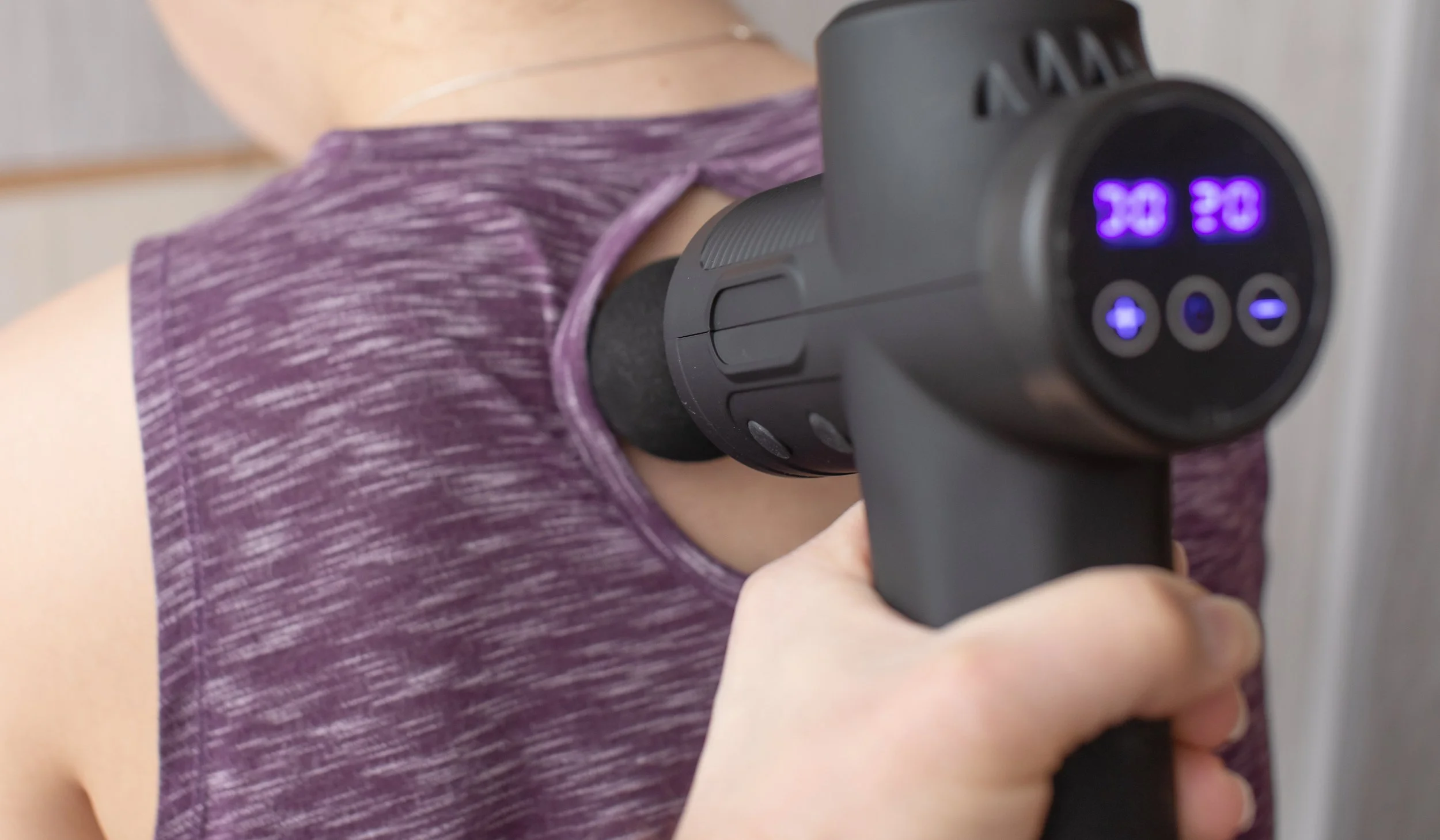 Person receiving a massage with a percussion massage gun on their shoulder, wearing a purple workout shirt.