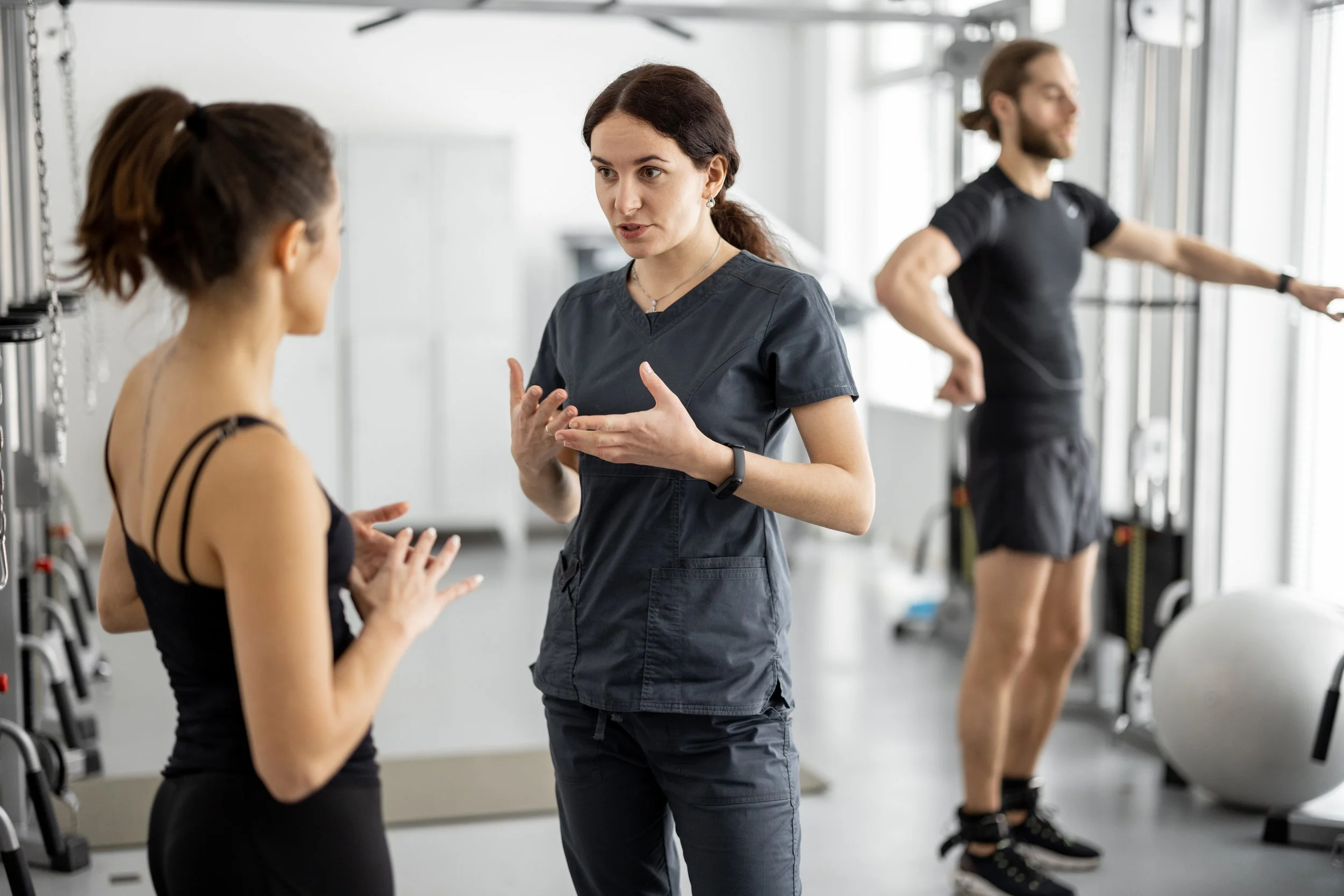 A female physical trainer talking with a woman gym member, while a male gym member exercises in the background in a bright gym.