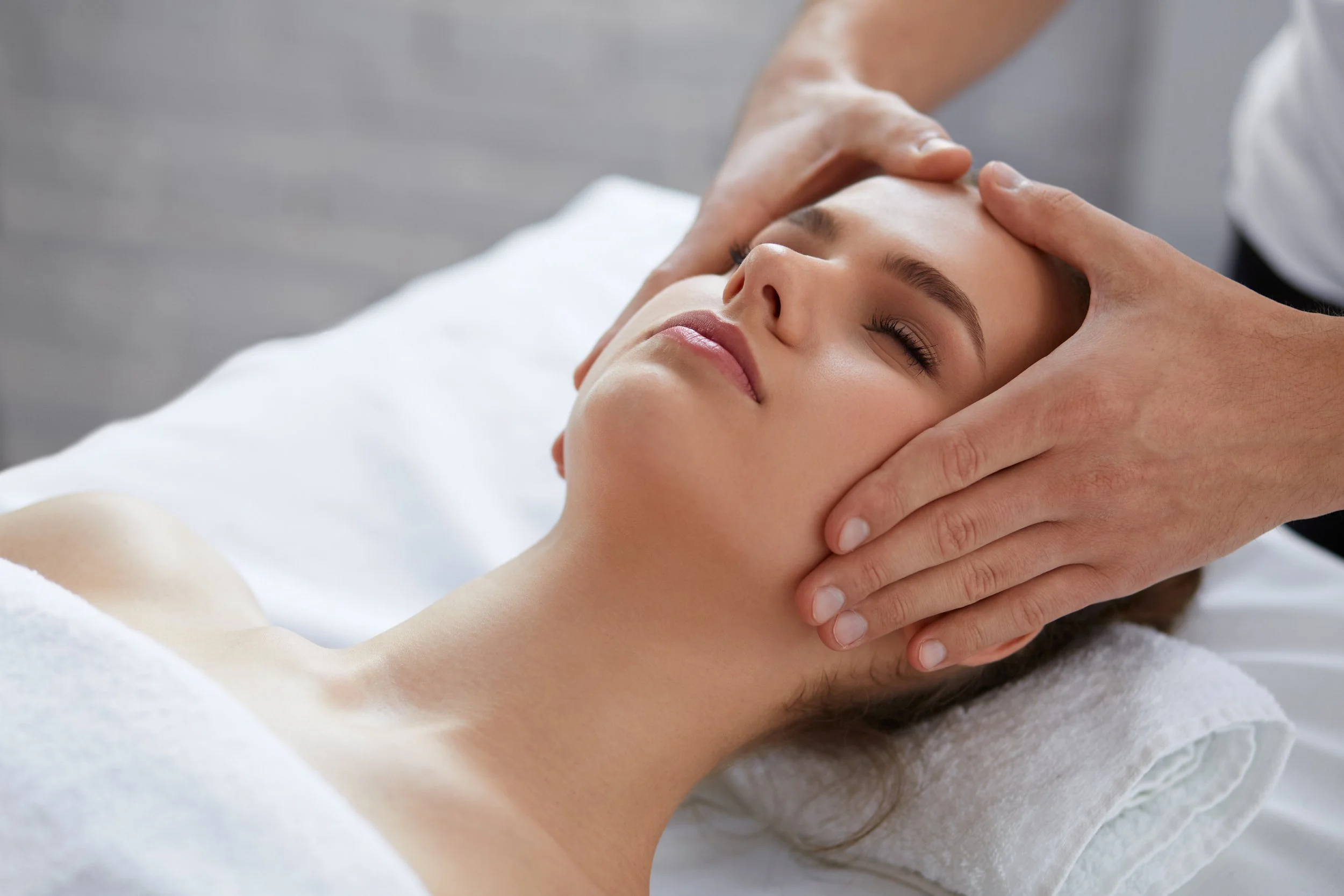 A woman receives a facial massage while lying on a treatment table with a white towel under her head.